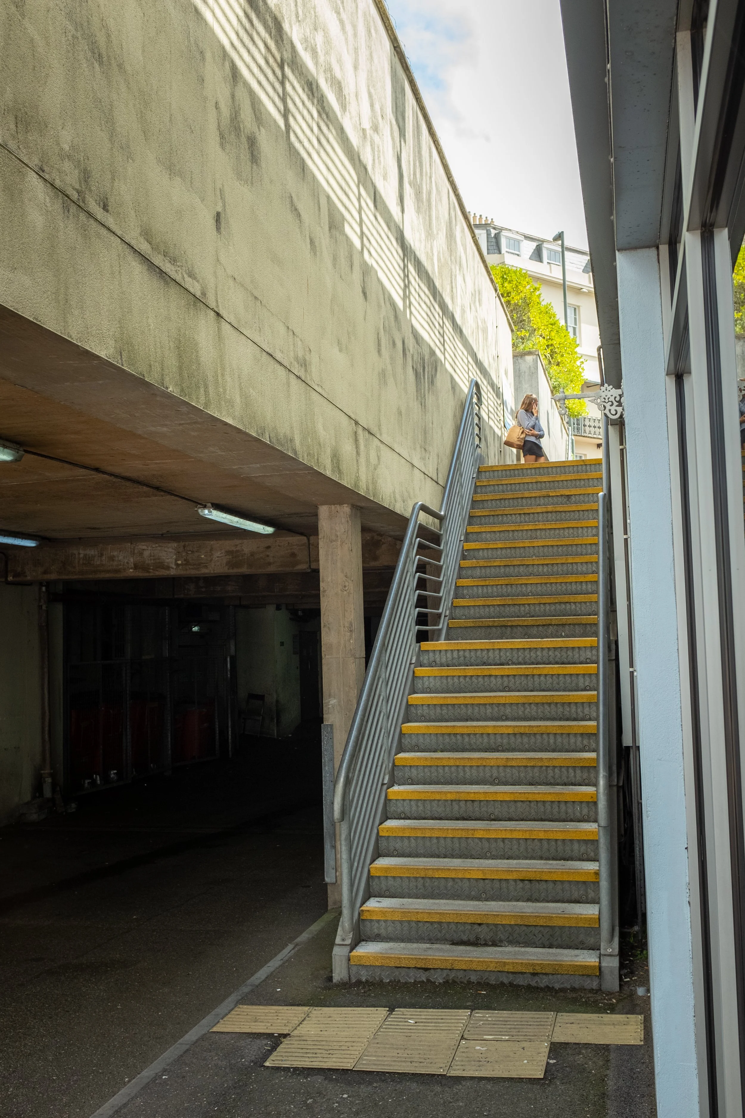 Outdoor staircase with yellow tripping strips leading up to a walk-way. A woman with brown hair, wearing a gray top, black shorts, and brown handbag, is standing at the top of the stairs looking at her phone. The scene is partly shaded with sunlight 