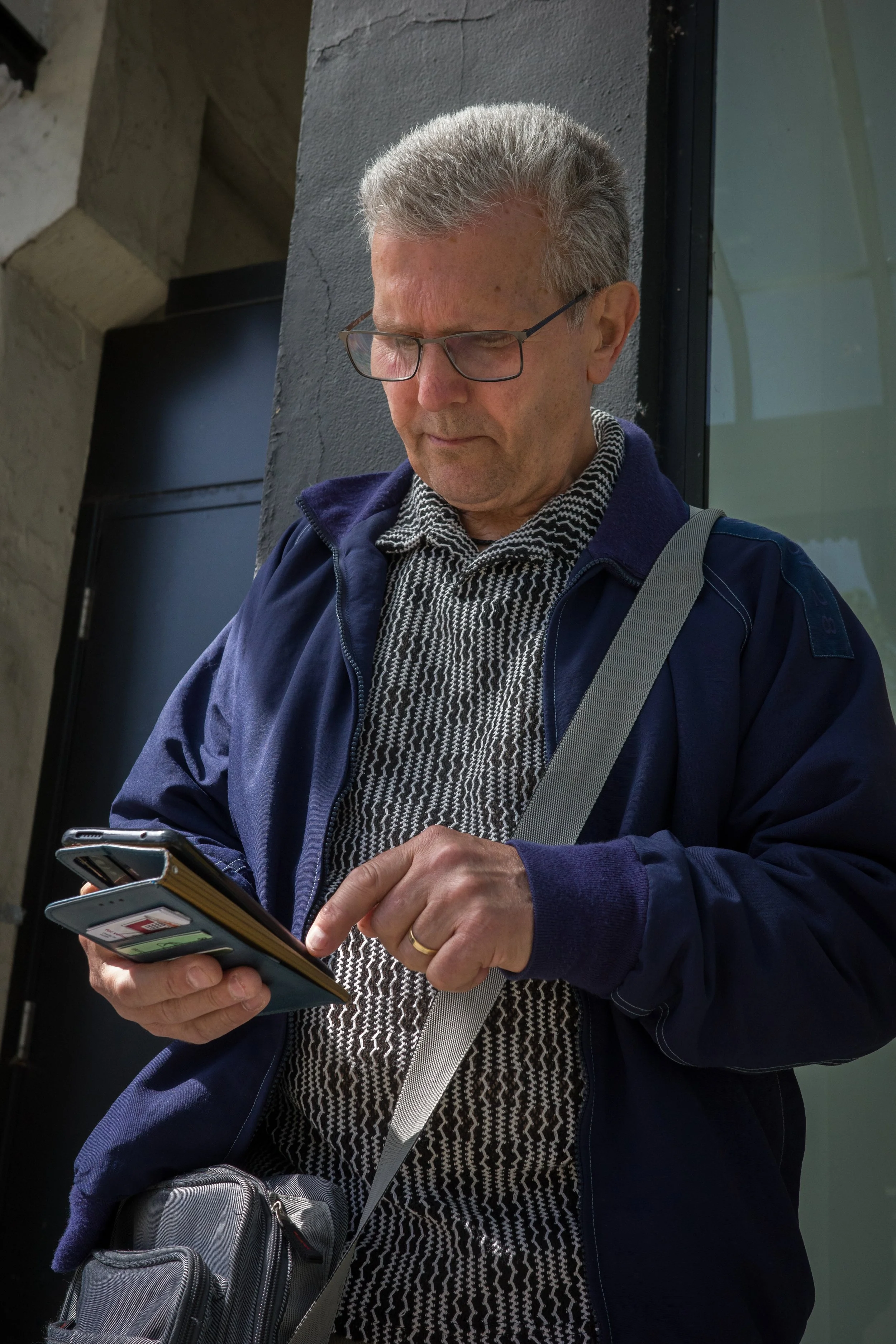 Man with gray hair and glasses looking at his phone outside, wearing a dark jacket and patterned shirt, carrying a gray bag over his shoulder.