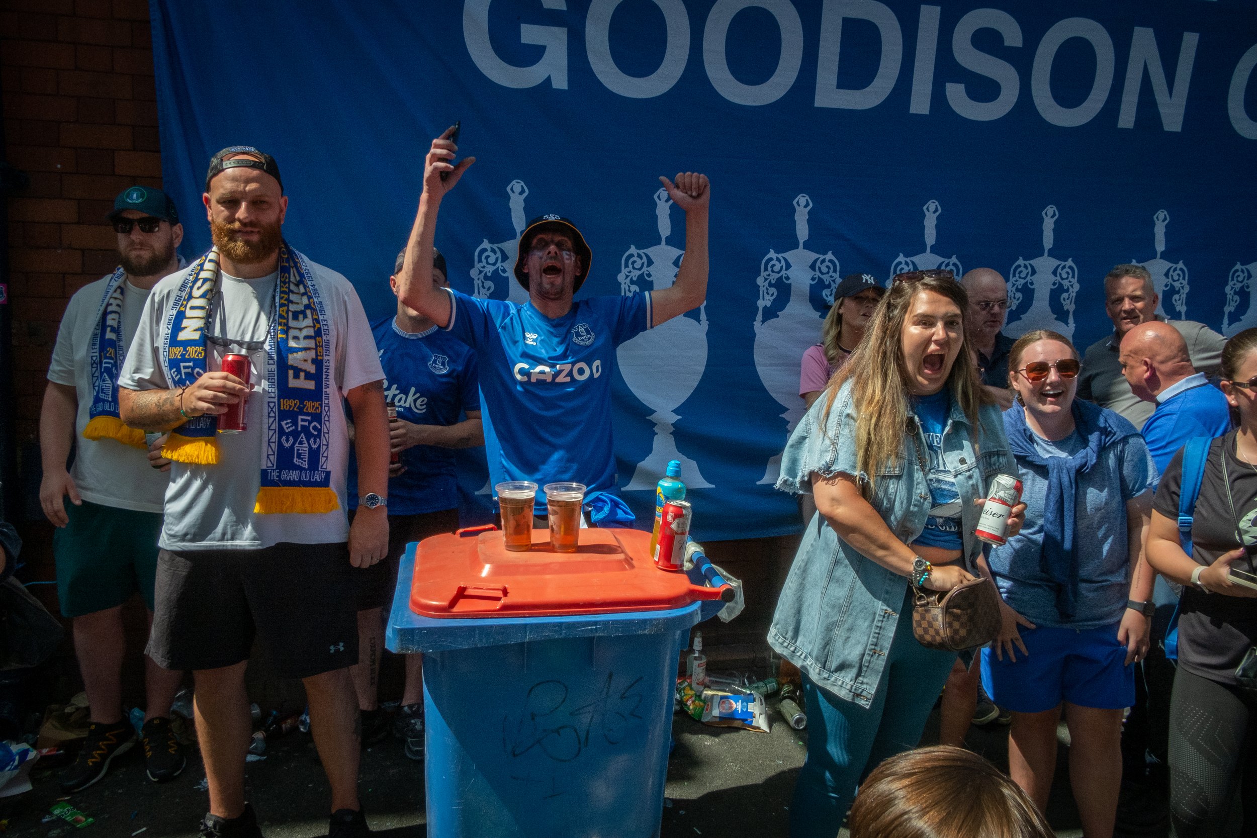 Group of Everton football fans celebrating in front of a blue banner that reads 'Goodison'. Some are wearing Everton team shirts and scarves, holding drinks, and cheering enthusiastically.