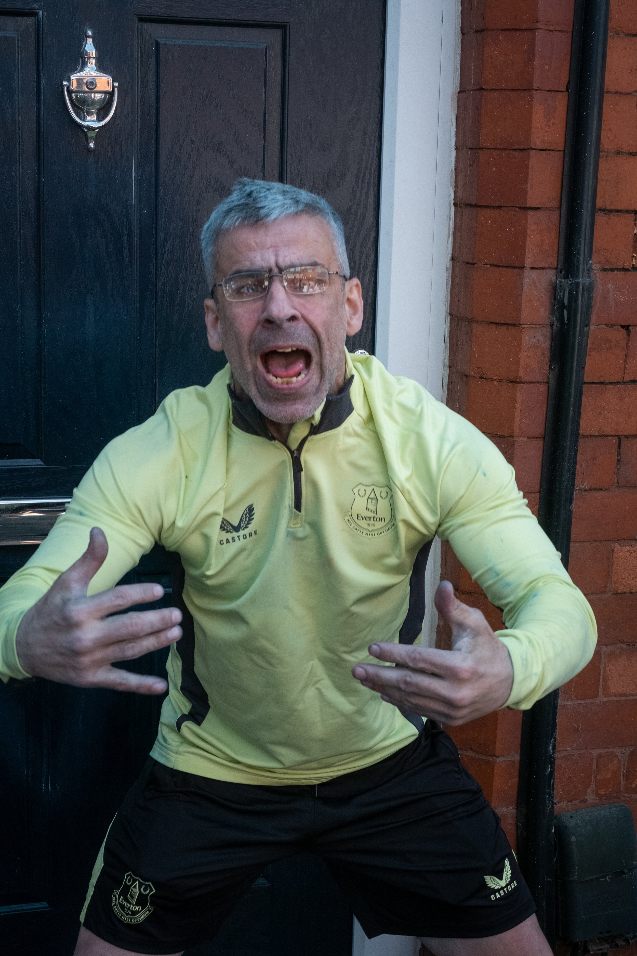 A man with gray hair and glasses wearing a yellow sports shirt and black shorts making an energetic gesture with his hands in front of a black door and brick wall.