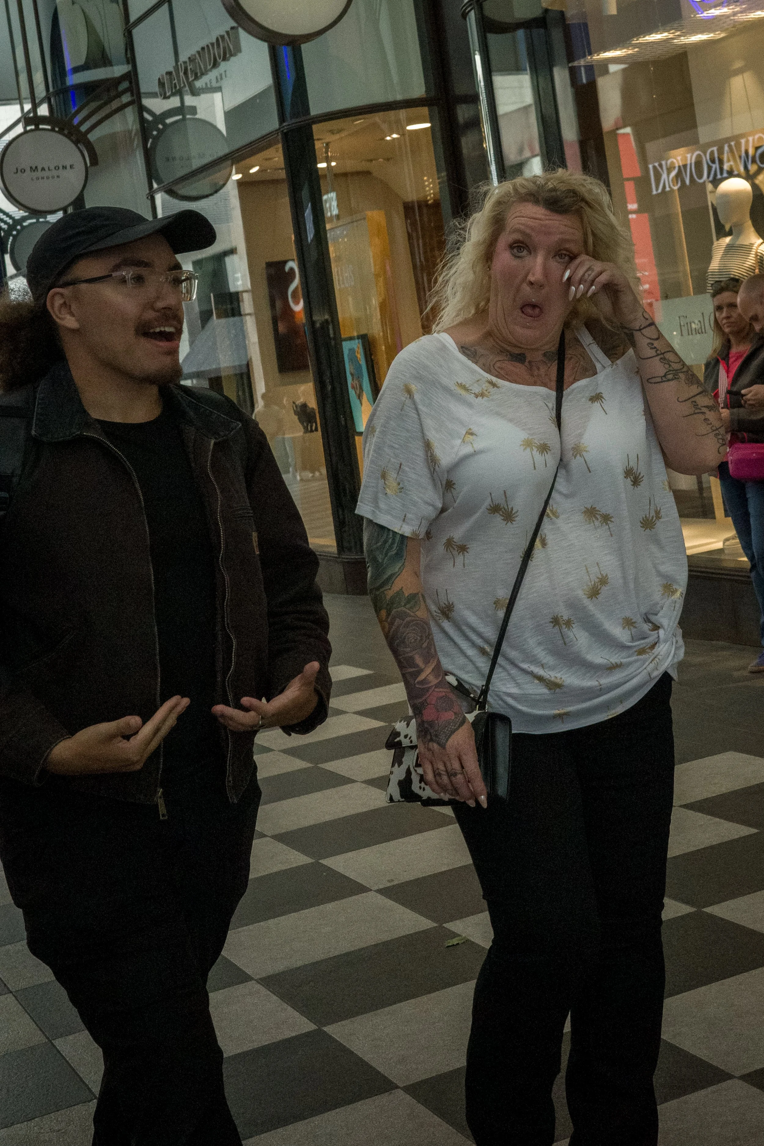 Two women walking in a shopping mall engaged in conversation; one is wearing a black jacket and cap, the other has blonde hair, tattoos, and is wearing a white shirt with palm tree prints.
