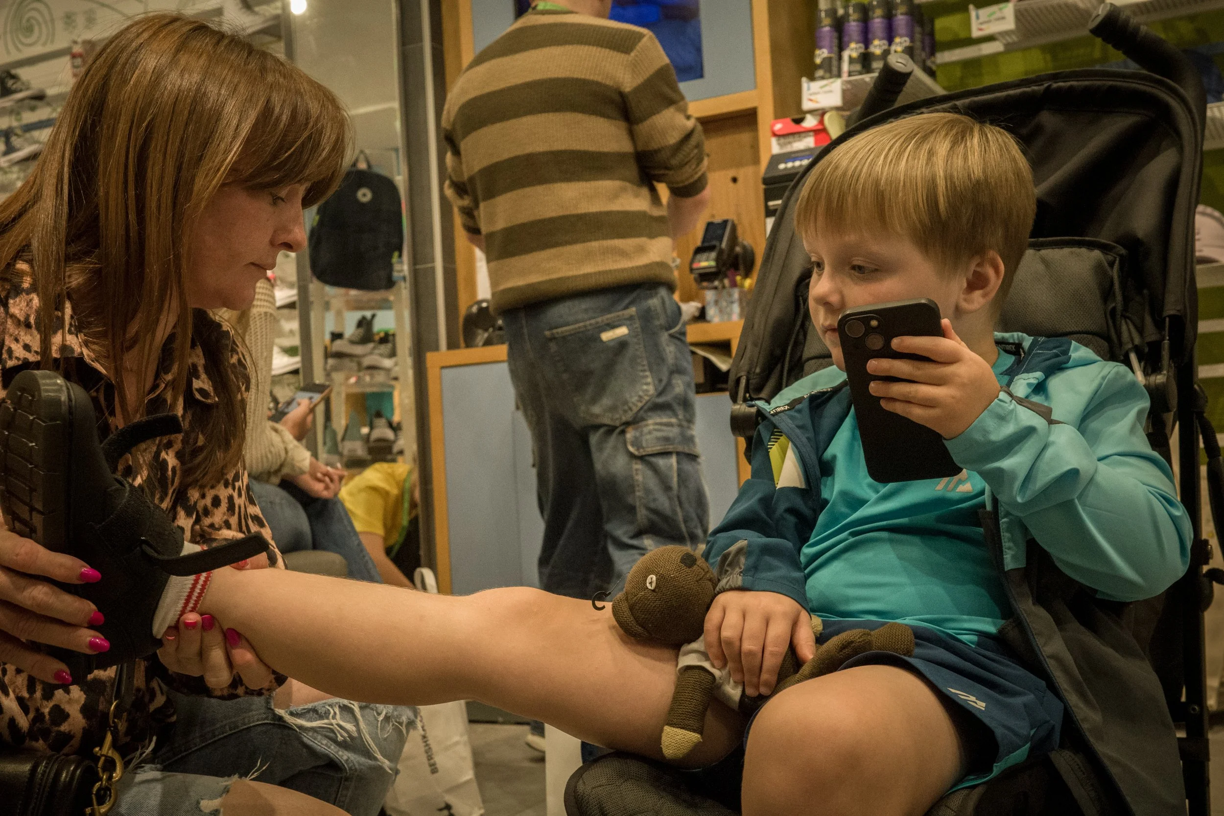 A woman with pink painted nails and a leopard print top is holding a young boy's leg up to examine it. The boy is sitting in a wheelchair, wearing a blue jacket, and looking at a smartphone. The woman appears to be inspecting the boy's leg, which has