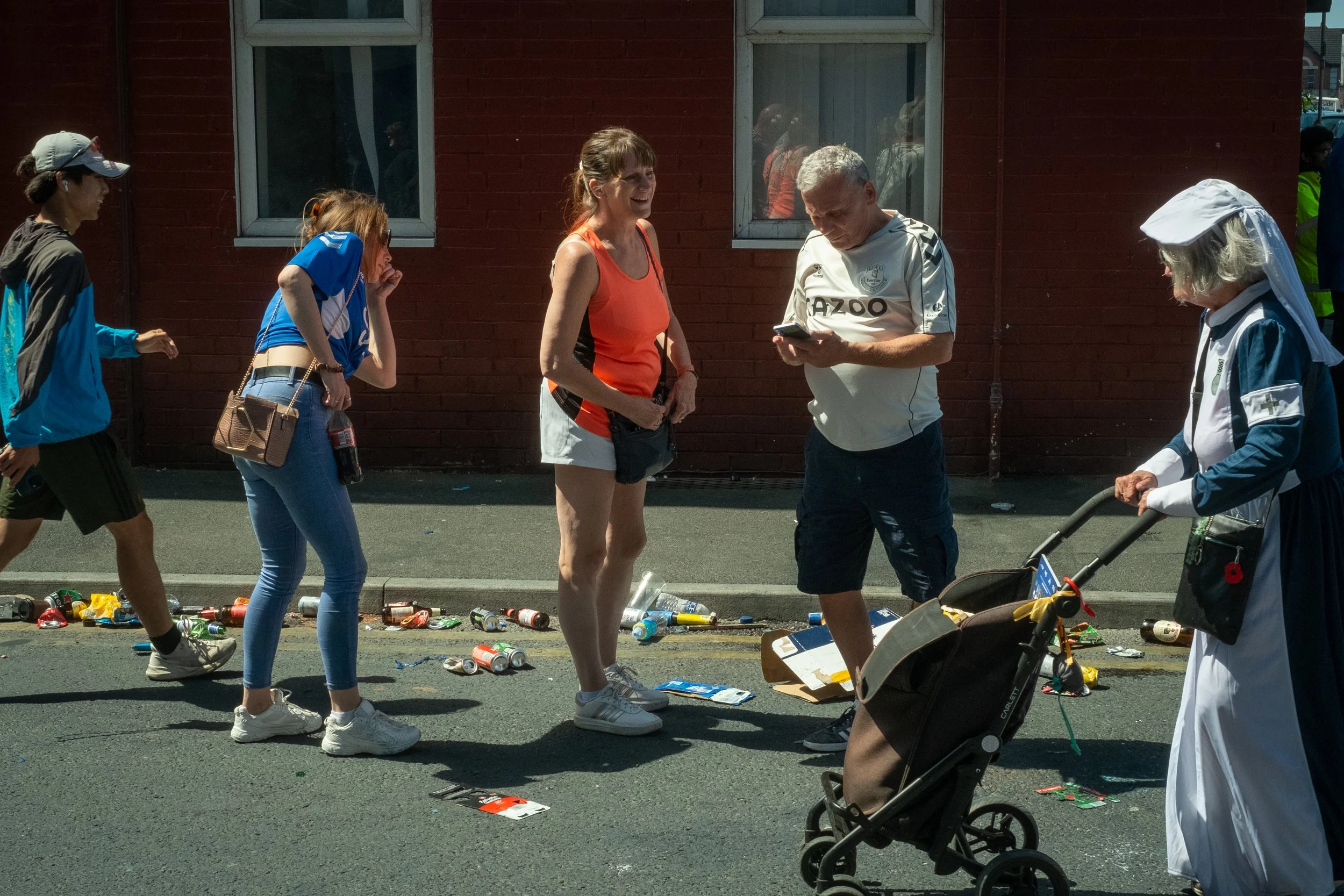 A group of people on a city street, with litter scattered on the ground, including cans and bottles, during daylight. Some juggle trash, others talk or walk, with a red brick building in the background.