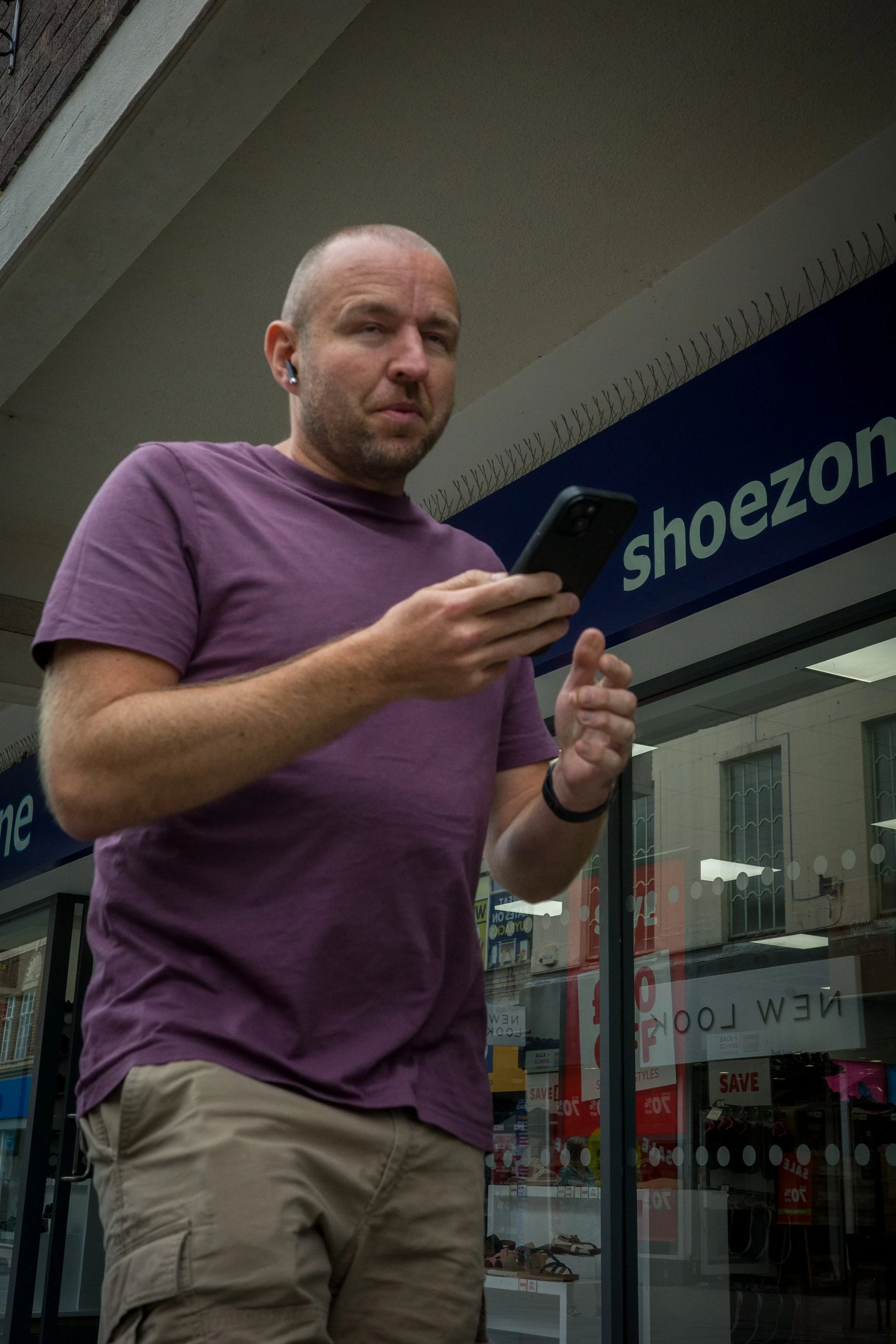 A man with a shaved head and beard wearing a purple t-shirt and beige cargo shorts standing outside a shoe store, looking at his phone.