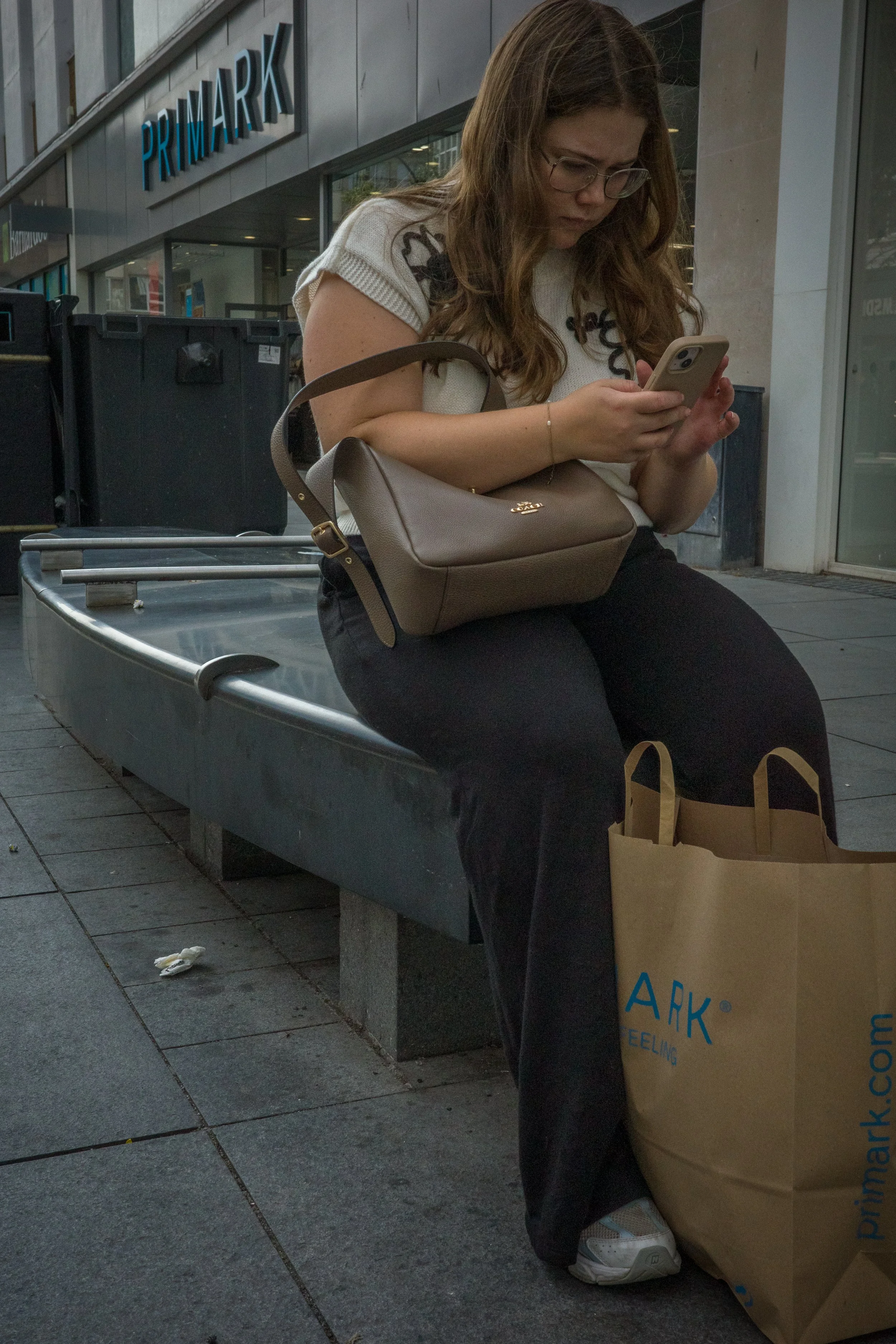 A woman sitting on a bench outside a Primark store, looking at her phone with a brown purse on her lap and shopping bags at her feet.