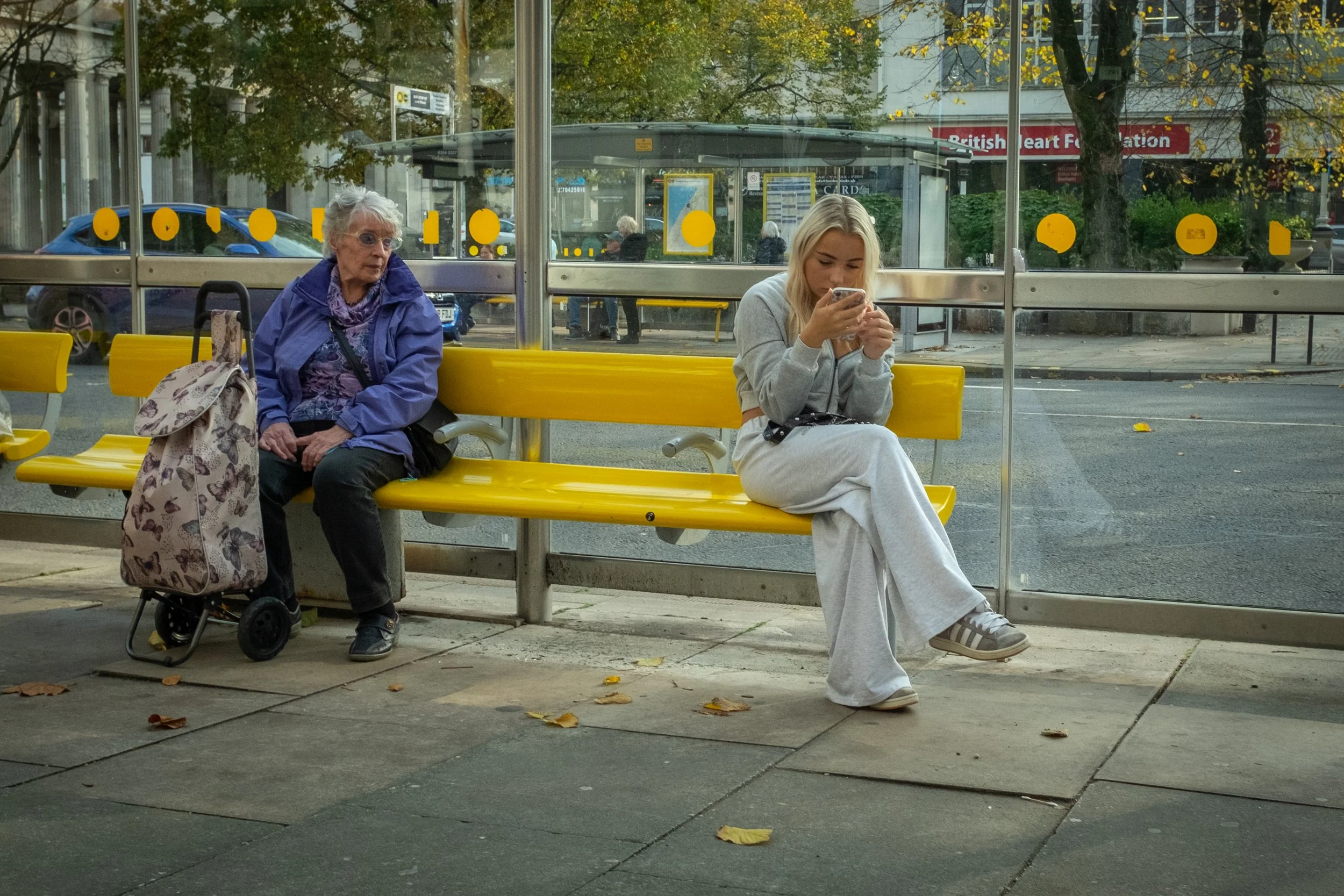 Two women sitting at a bus stop on yellow benches, with a glass shelter behind them. An elderly woman with a mobility cart and a bag sits on the left, wearing a purple jacket and reading glasses. A young woman with long blond hair, dressed in a gray 