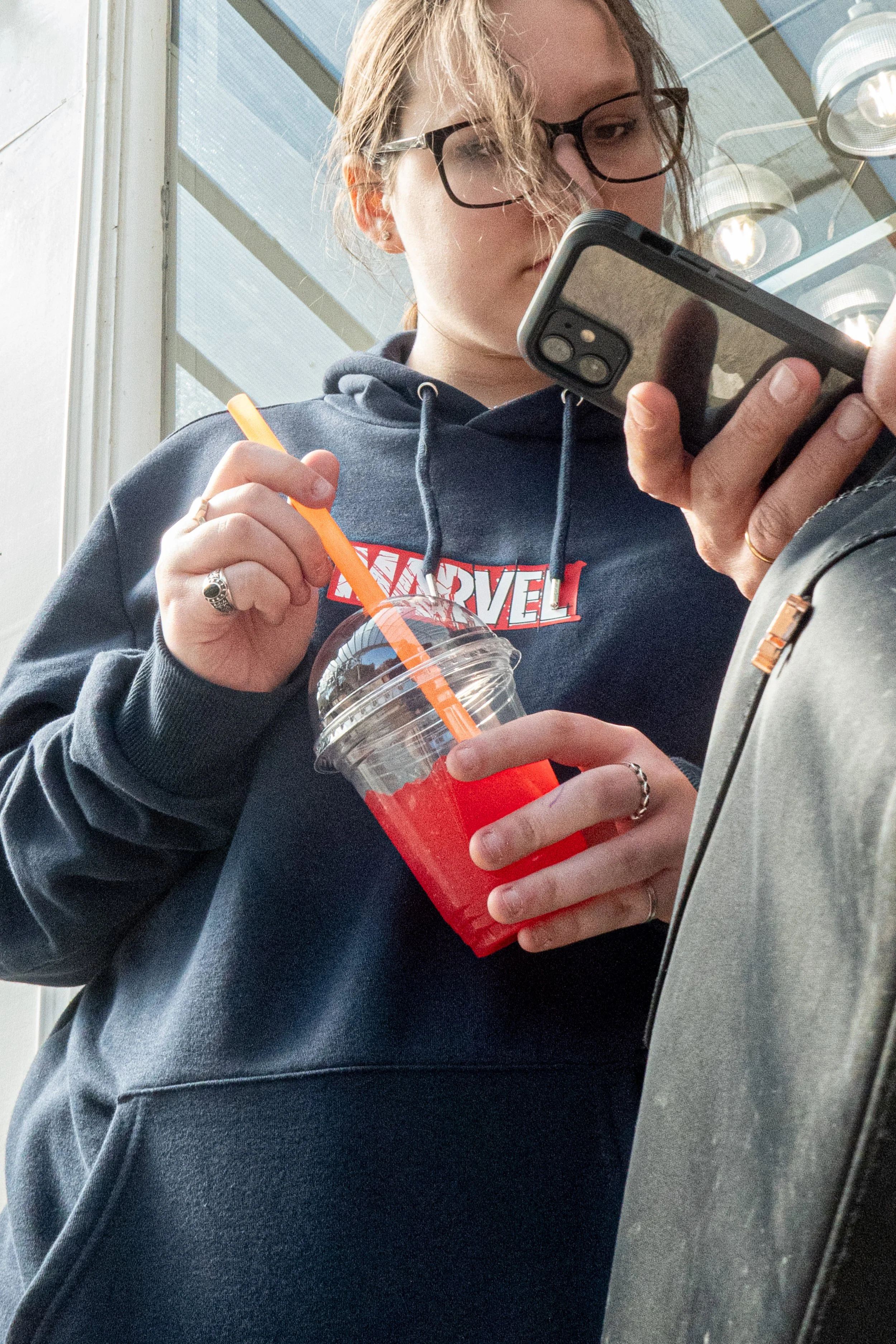 A woman with glasses wearing a navy hoodie with a Marvel logo, holding a plastic cup with a red beverage and drinking through an orange straw, standing indoors near a window.