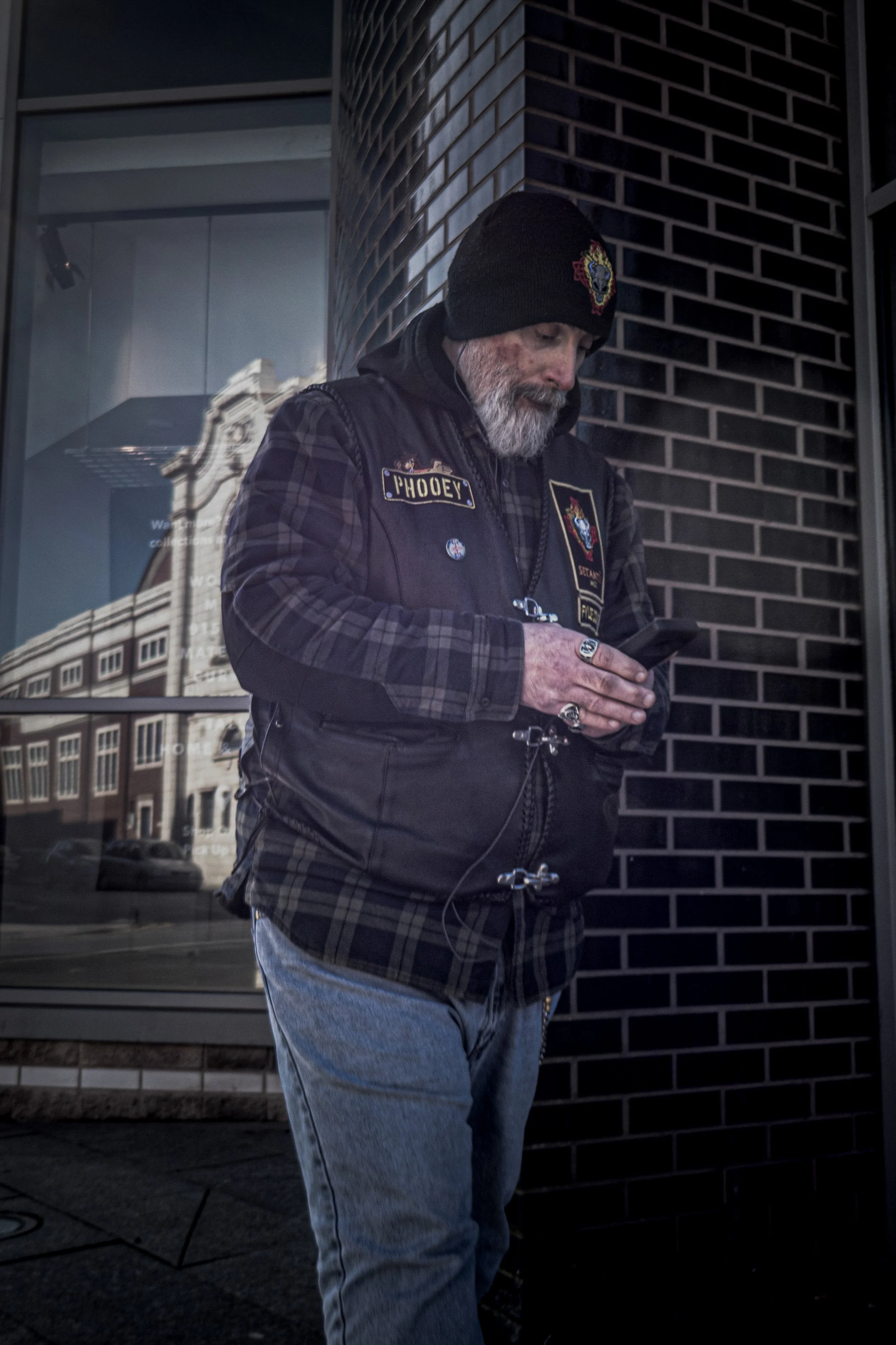 A man with a gray beard wearing a black beanie, a black vest with patches, and blue jeans, standing outside near a brick wall, looking at his phone.