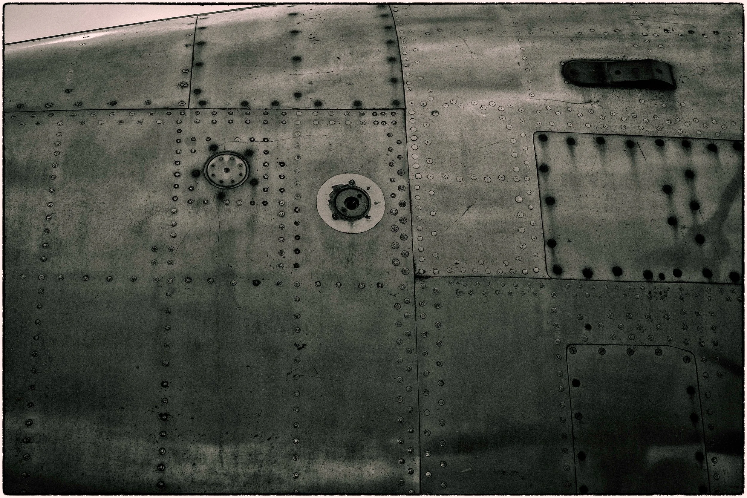 Close-up of a metallic surface, likely part of an aircraft fuselage, with numerous rivets and two circular hatch covers, one open and one closed, and a small rectangular access panel.
