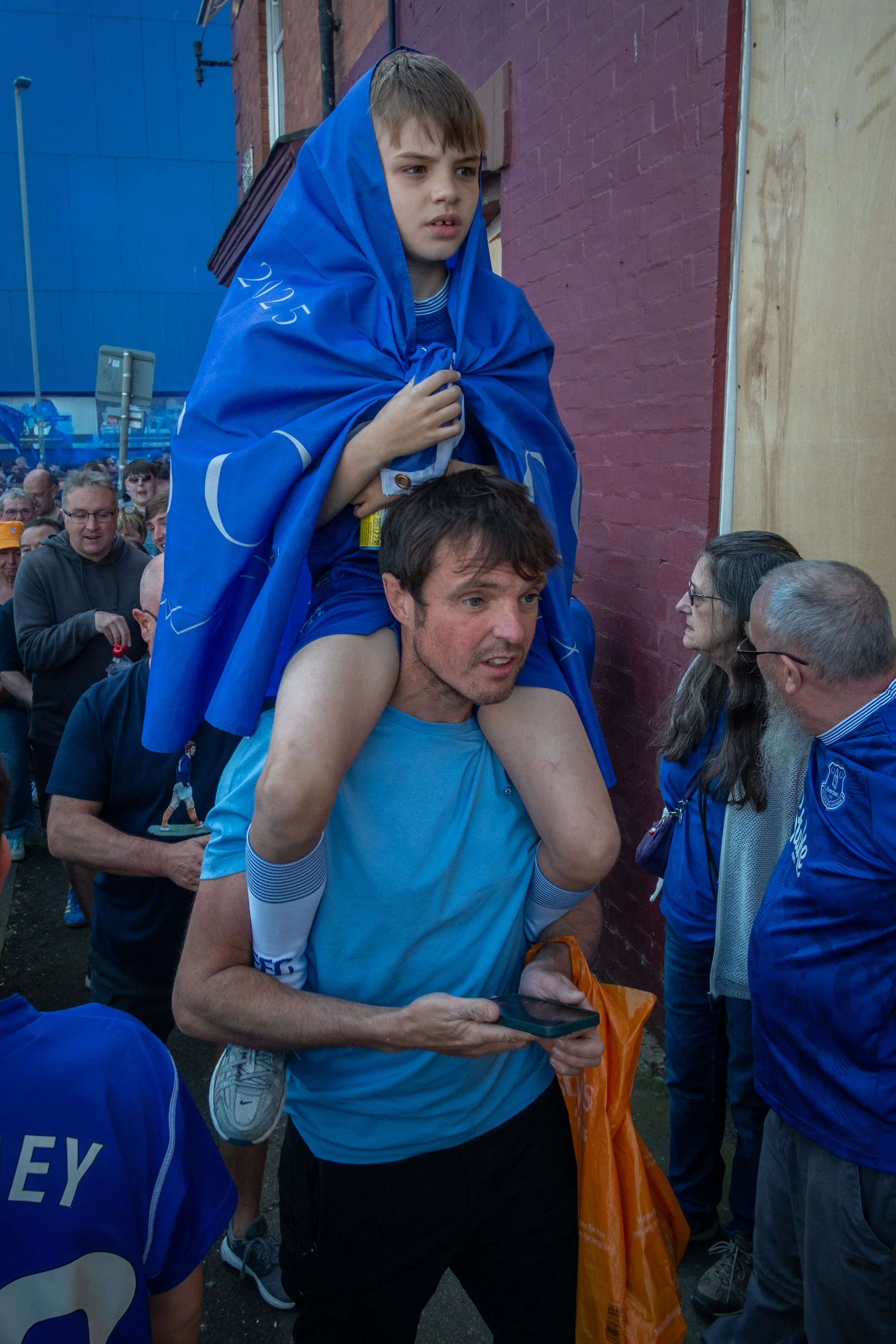 A man carries a young boy on his shoulders, surrounded by a crowd at an outdoor event. The boy is wearing a blue cloak with '2015' printed on it, and the man is holding a smartphone.