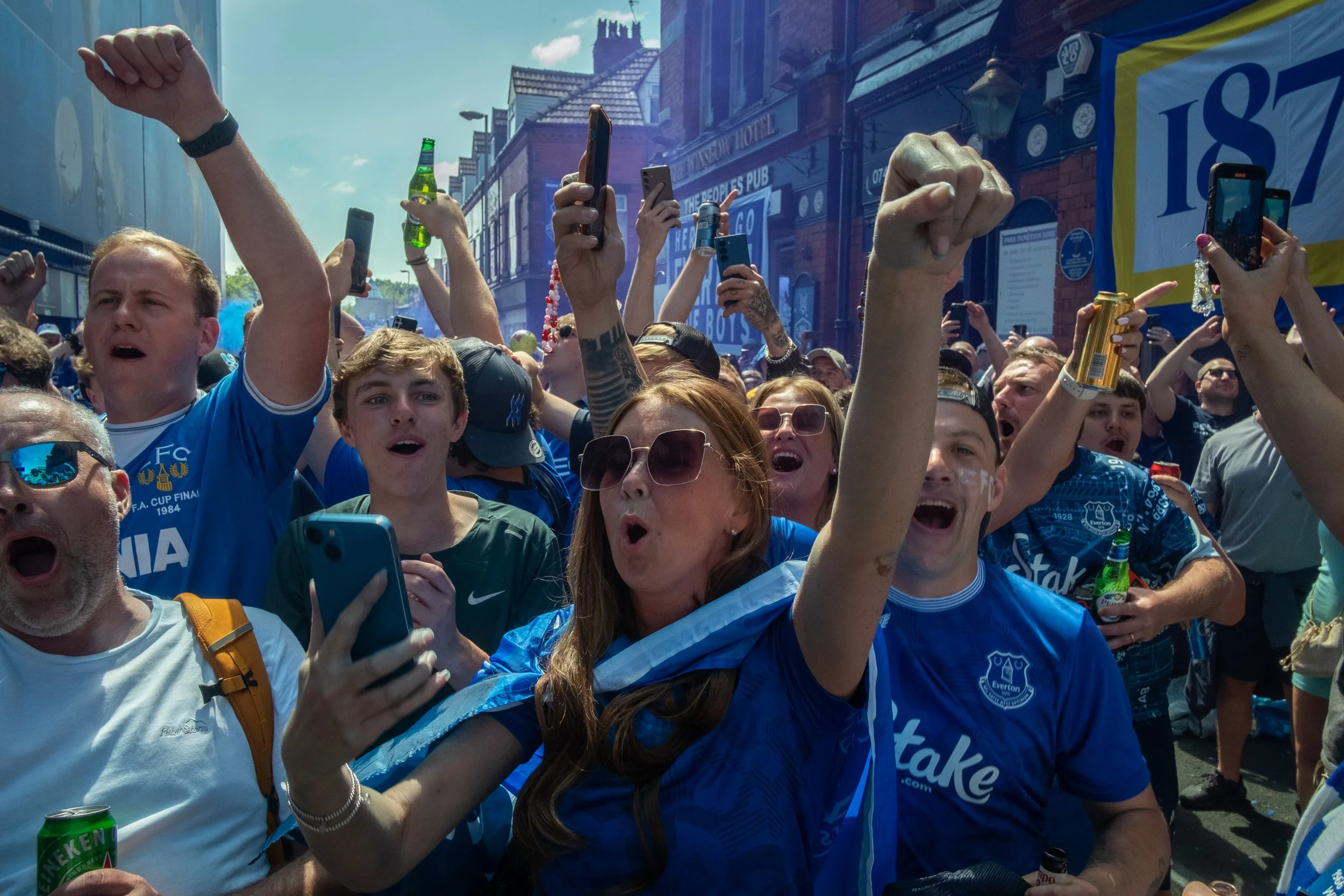 Crowd of football fans celebrating outdoors, many wearing blue jerseys, some wearing sunglasses, cheering and taking photos.