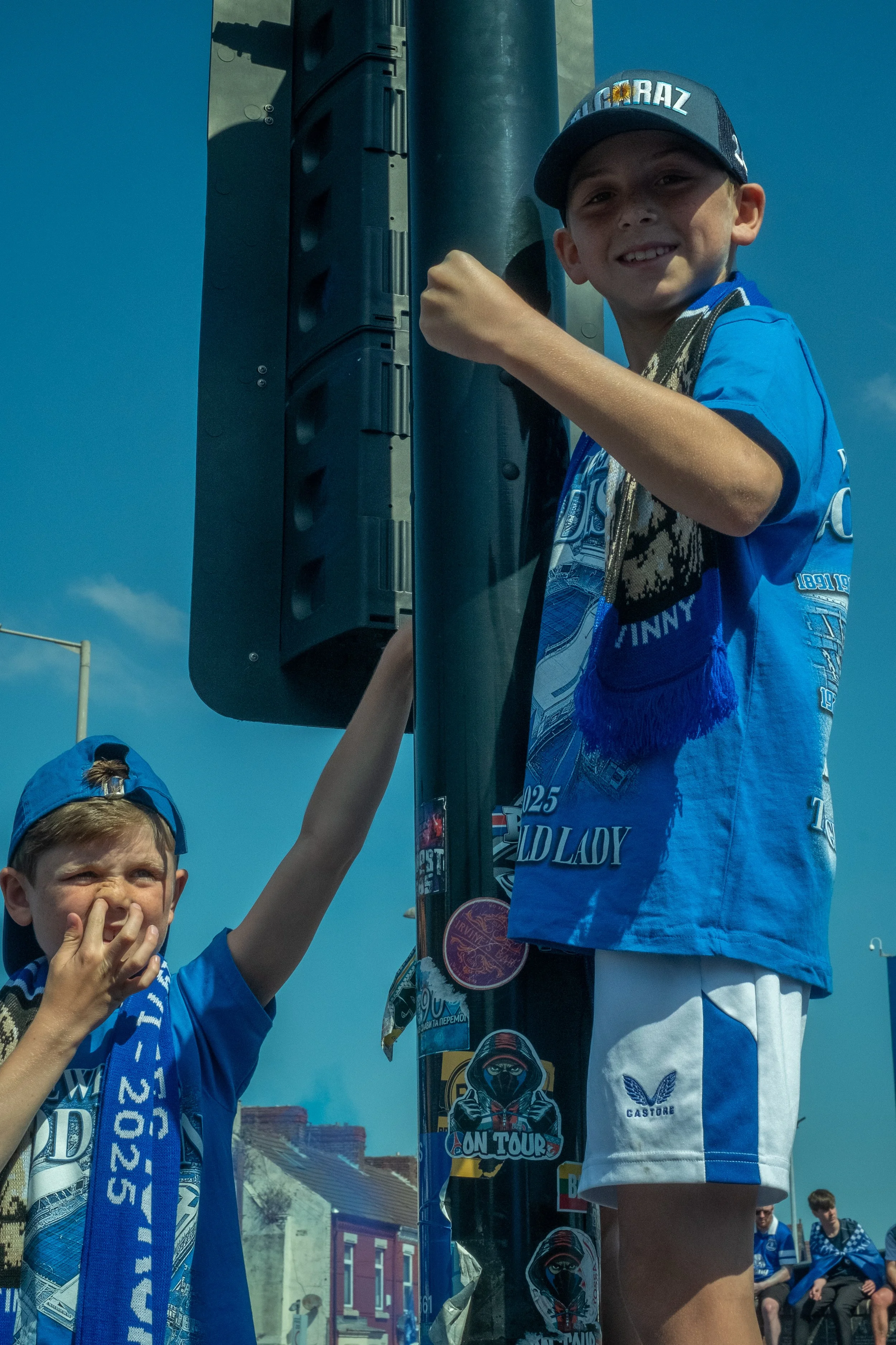 Two young boys wearing blue t-shirts and hats standing next to a streetlight with stickers on it, one of them smiling and woman standing next to the streetlight. The woman is wearing a blue t-shirt and a cap, smiling at the camera. The sky is clear a