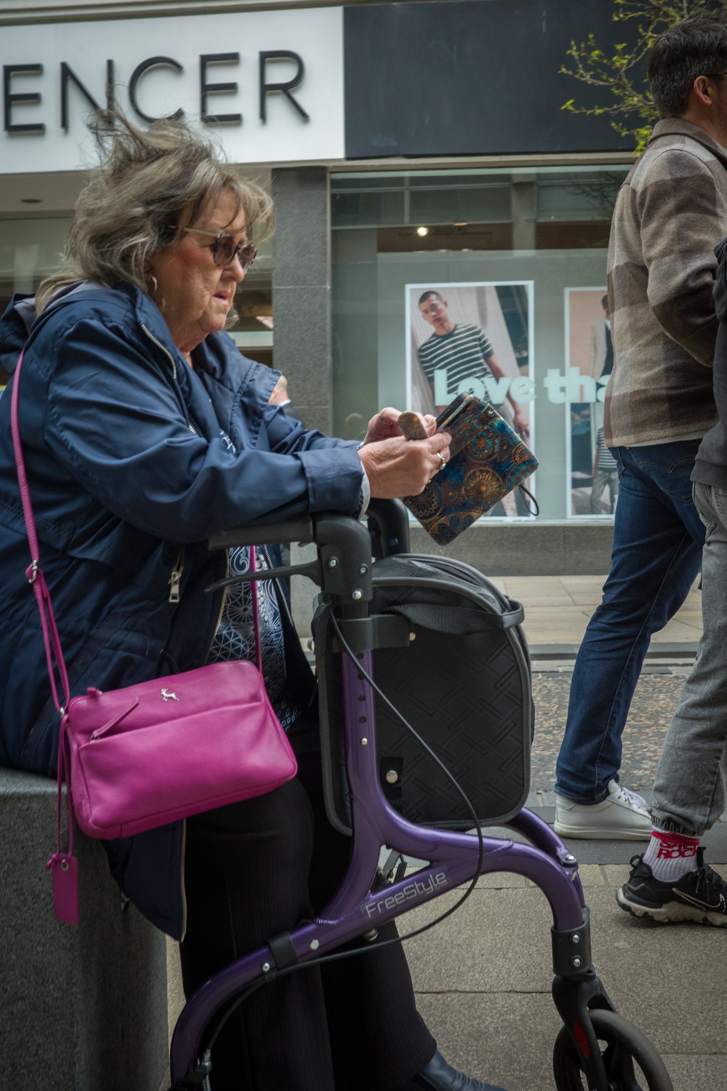 A woman with gray hair, glasses, and a blue jacket sitting on a bench with an electric purple scooter in front of her, holding a phone and a patterned bag, with a pink purse hanging on the scooter, in an urban setting.