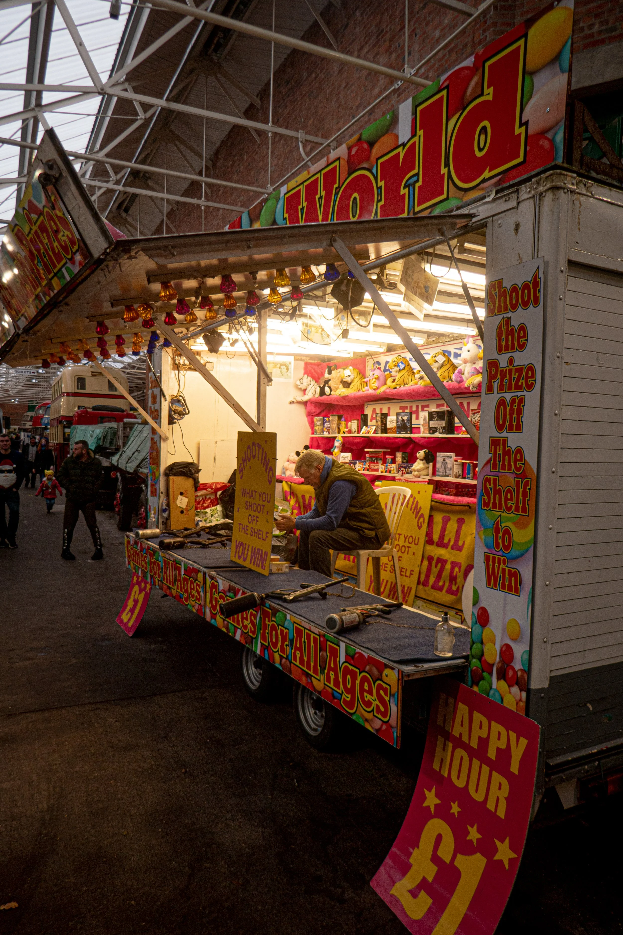 A carnival game booth with bright, colorful signage and stuffed toys for prizes, under a metal roof, with people walking nearby.