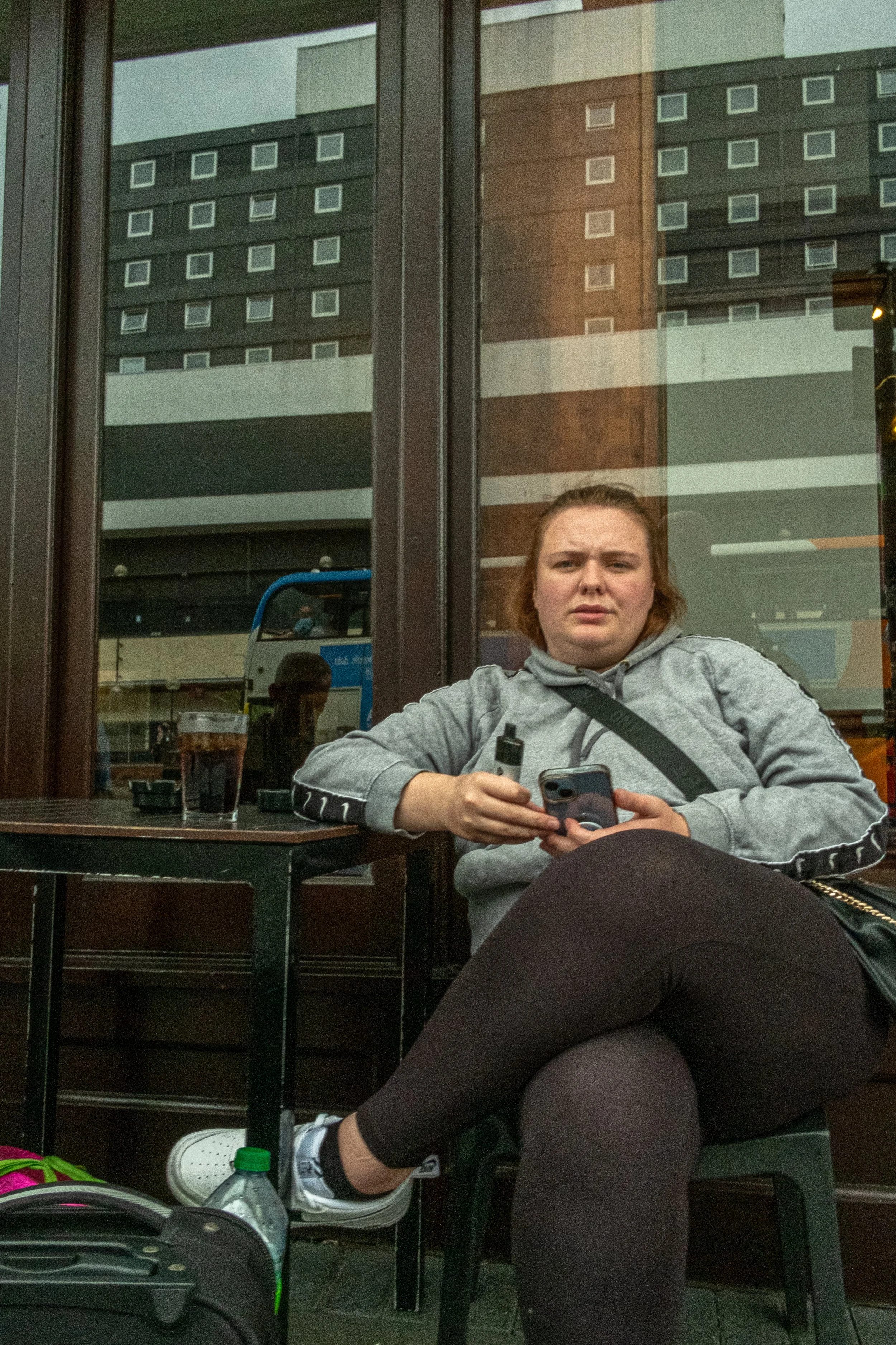A woman with red hair and a gray hoodie is sitting outside a café or restaurant, holding a smartphone. She appears to be looking at the camera with a slightly confused or surprised expression. She's sitting at a small table with a drink in front of h