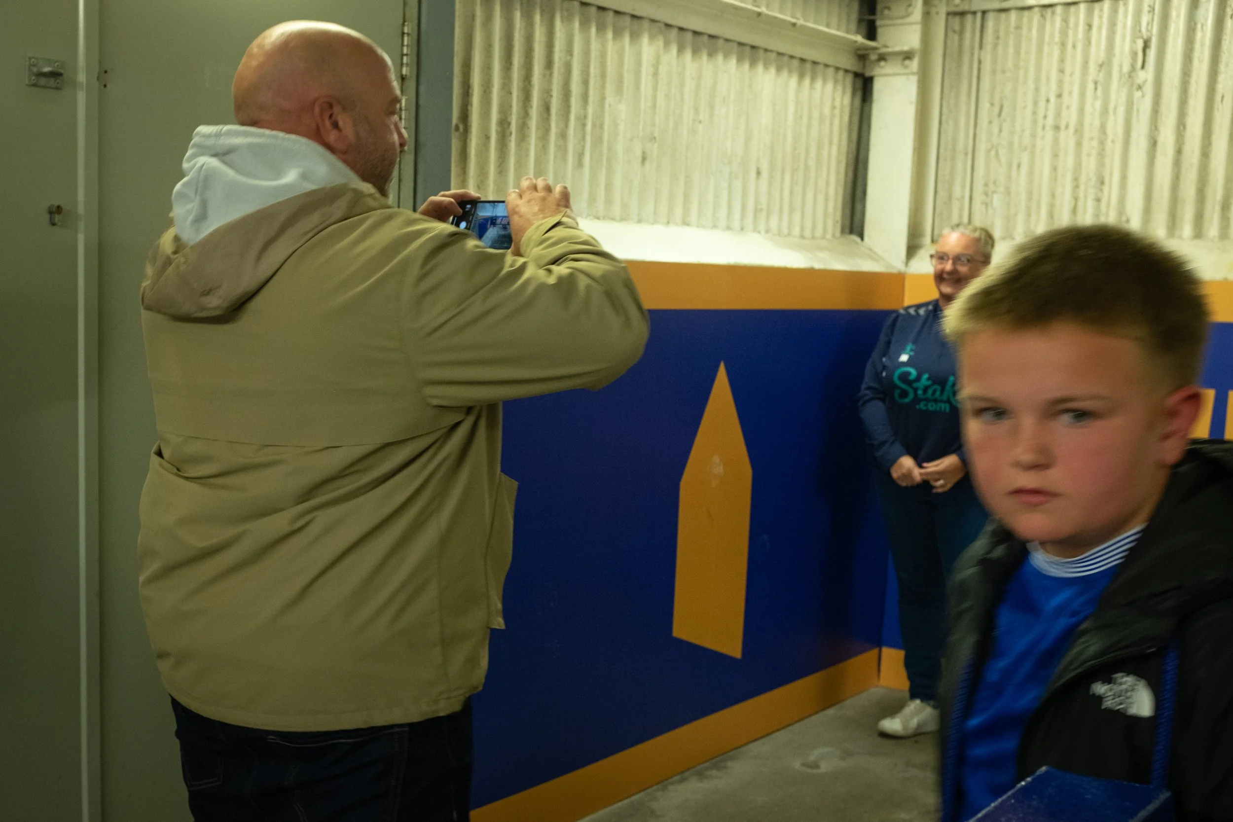 A man with a bald head and beige jacket is taking a photo of a smiling woman with glasses, who is standing next to a child, in an indoor sports facility with a blue and yellow wall and wooden panels.