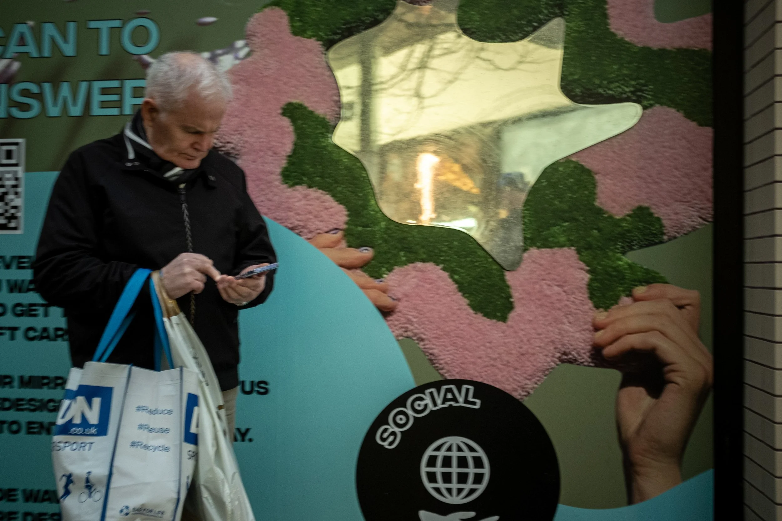 A person with white hair looking at their phone, carrying a tote bag with recycling hashtags, standing in front of an environmental-themed poster with hands and a puzzle piece design.