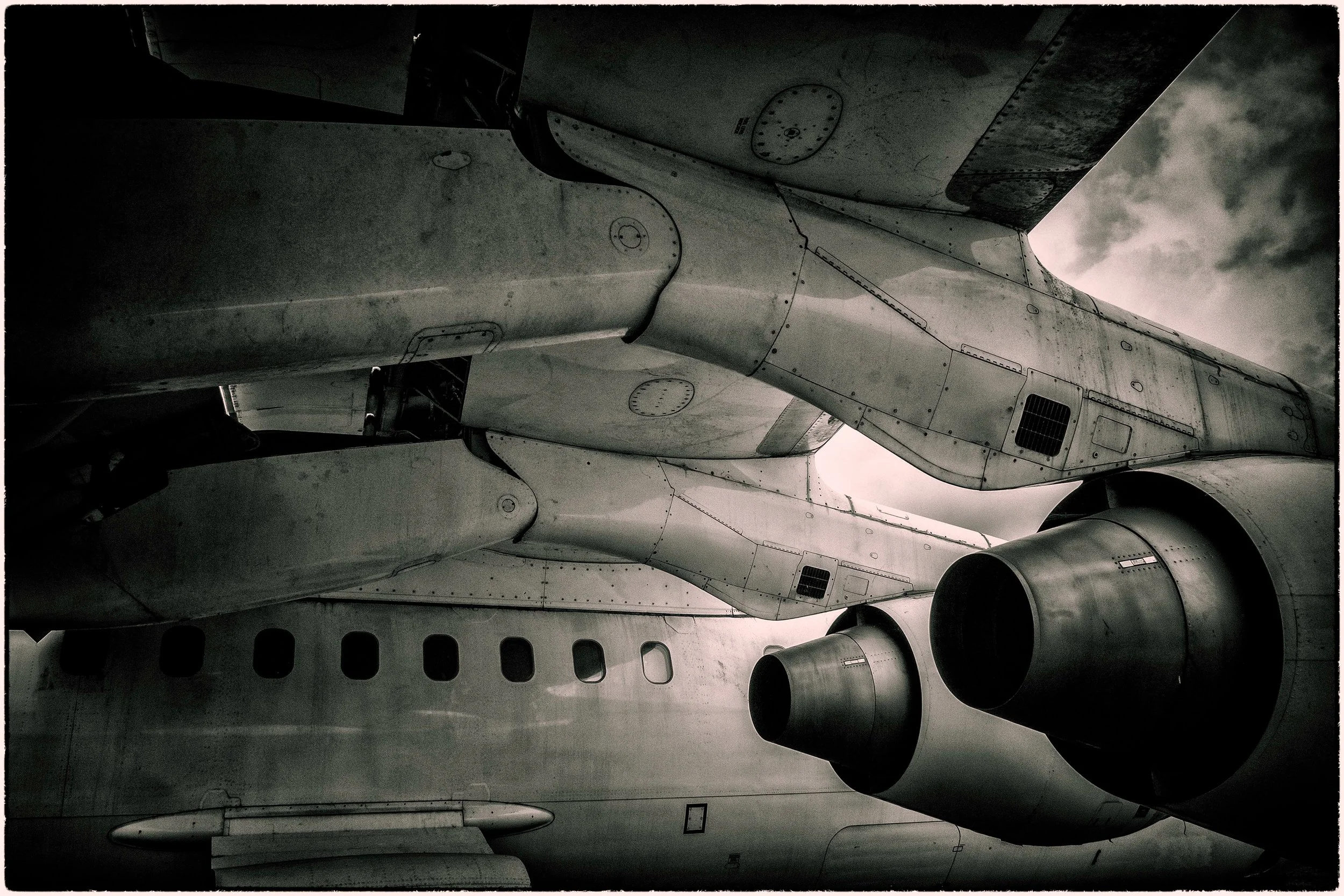Close-up view of the underside of a fighter jet aircraft, showing parts of the wing, engines, and fuselage with cloudy sky in the background.