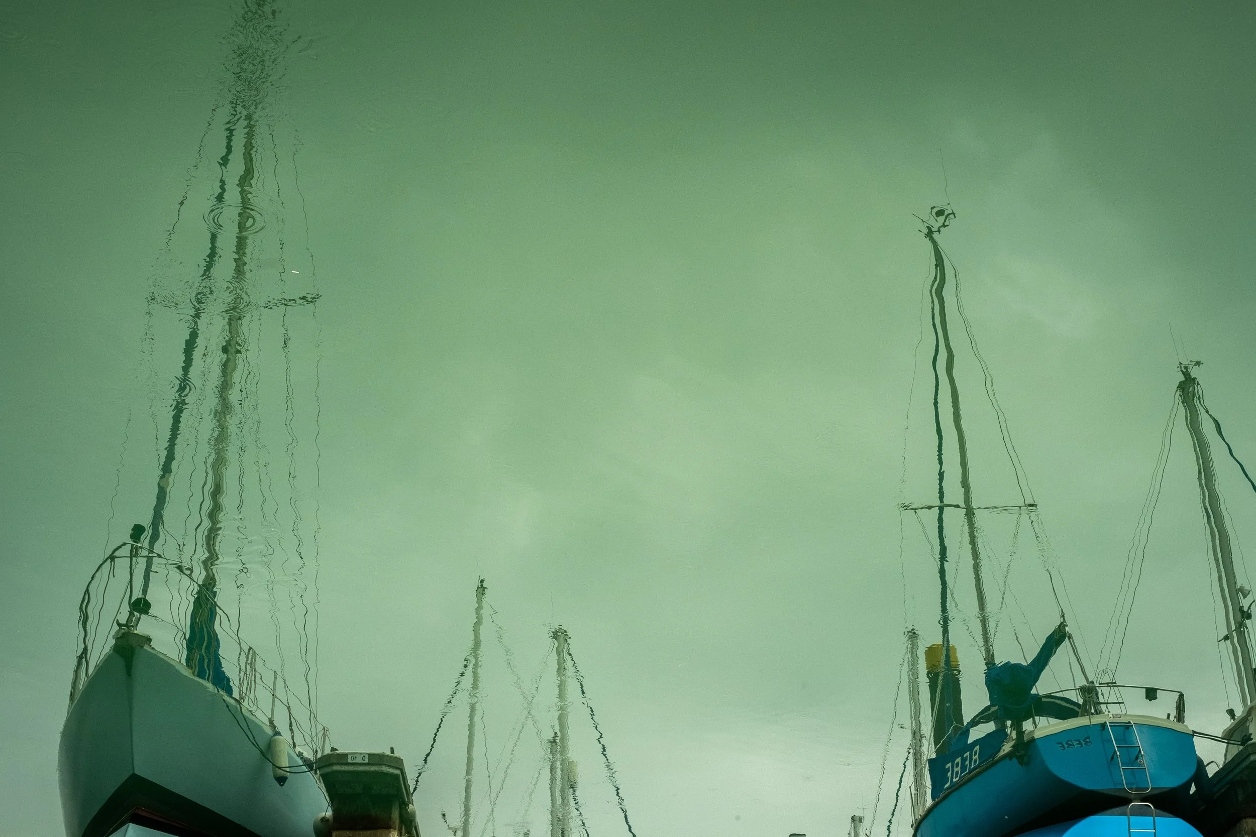 Reflections of sailboats in greenish water, with tall masts and rigging visible.
