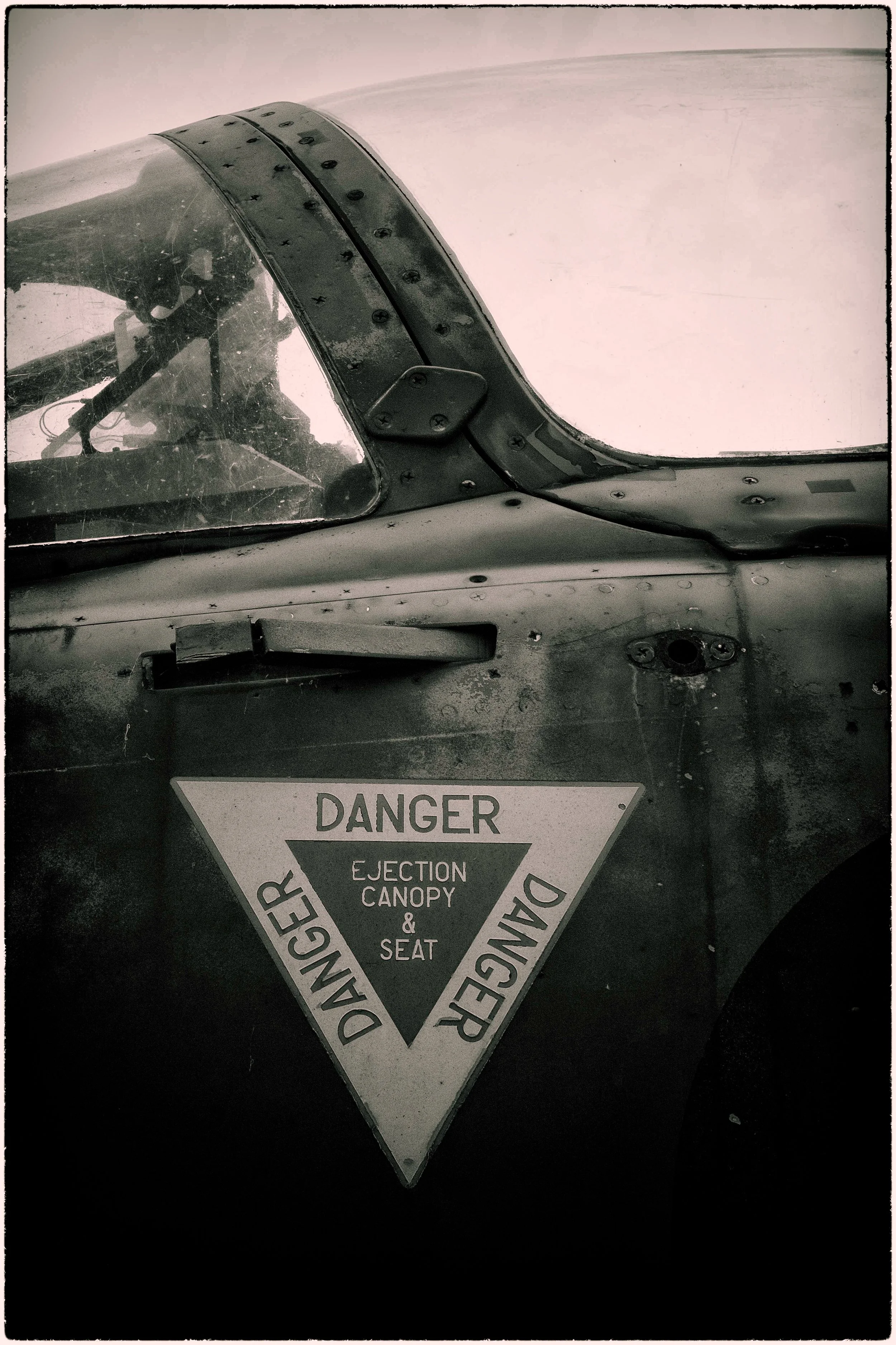 Close-up of a worn warning sign on an aircraft indicating danger due to ejection canopy and seat, with visible screws and surface scratches.