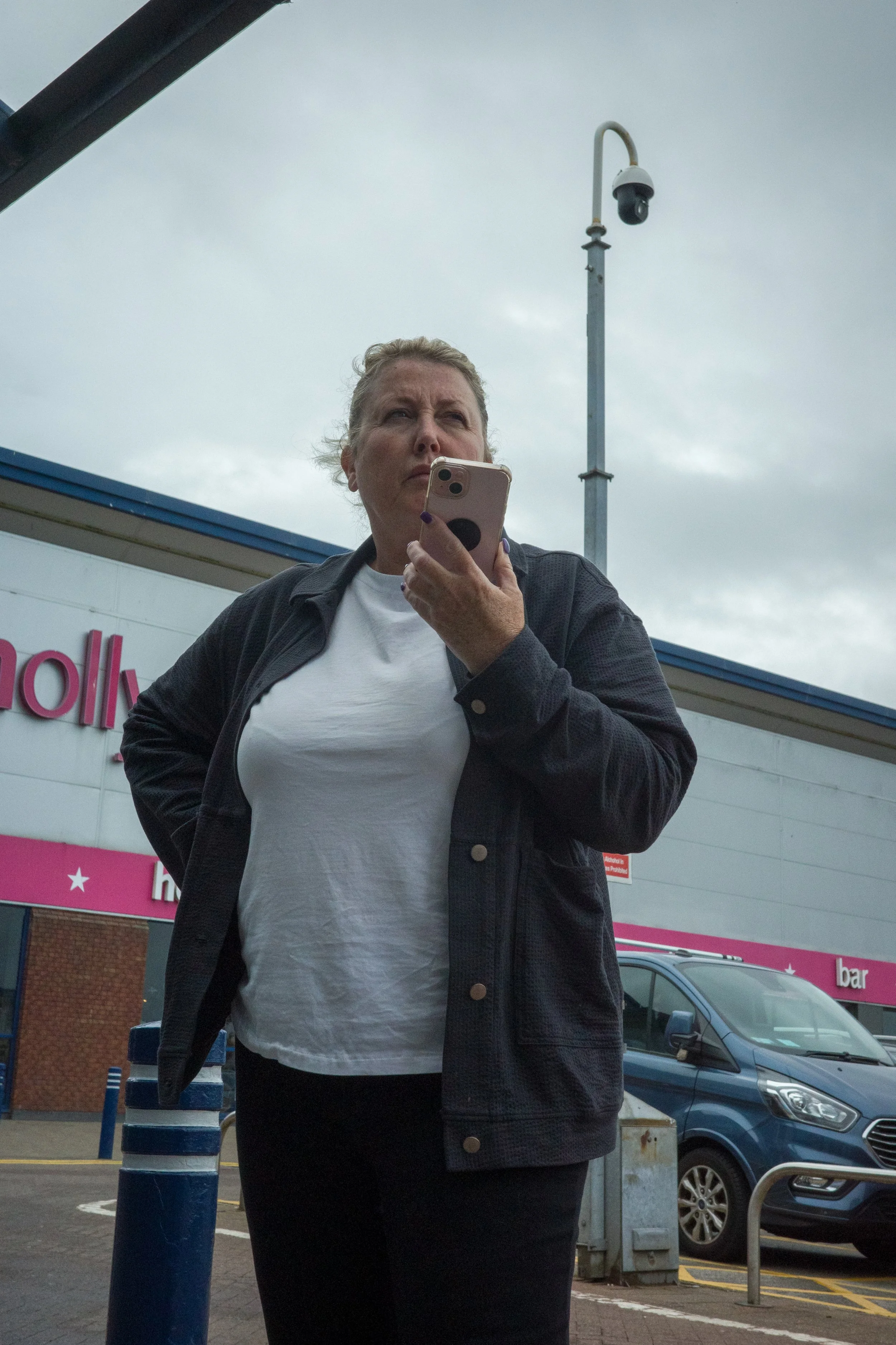 A woman standing outside a shopping center, holding a smartphone, with dark clouds overhead, cars parked nearby, and a purple and pink store sign partially visible in the background.