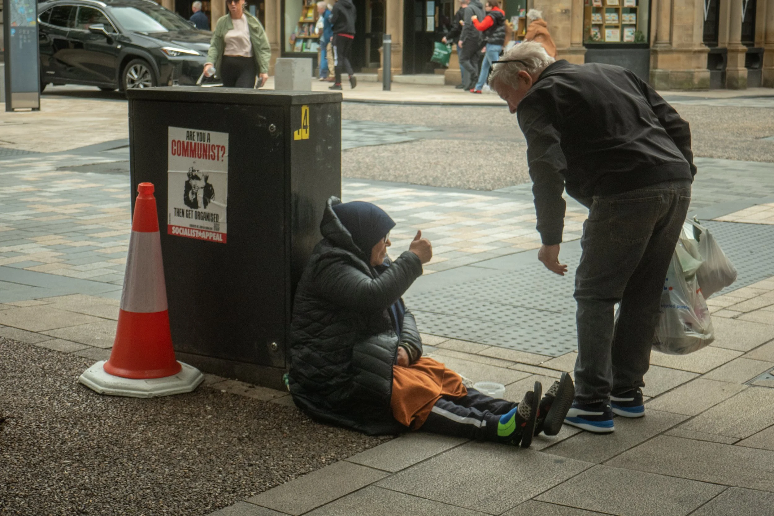 A person sitting on the sidewalk next to a black utility box, giving a thumbs-up to another person standing nearby. The person sitting is wearing a black jacket, a beanie, and orange pants. The person standing is wearing a black jacket and jeans, hol