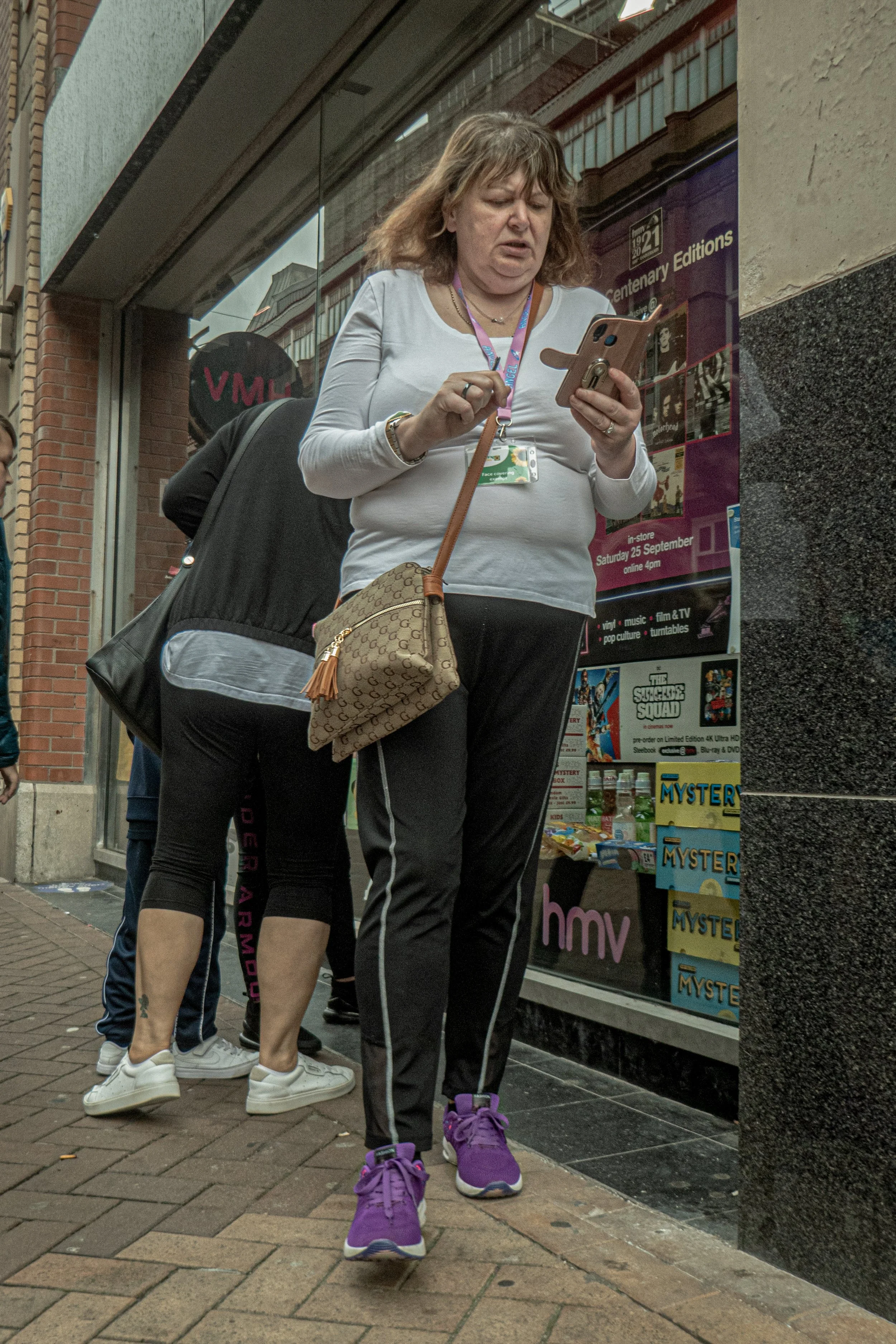 A woman in athletic clothing and purple sneakers stands on a sidewalk looking at her phone outside a store with a display of DVDs and a sign for HMV.