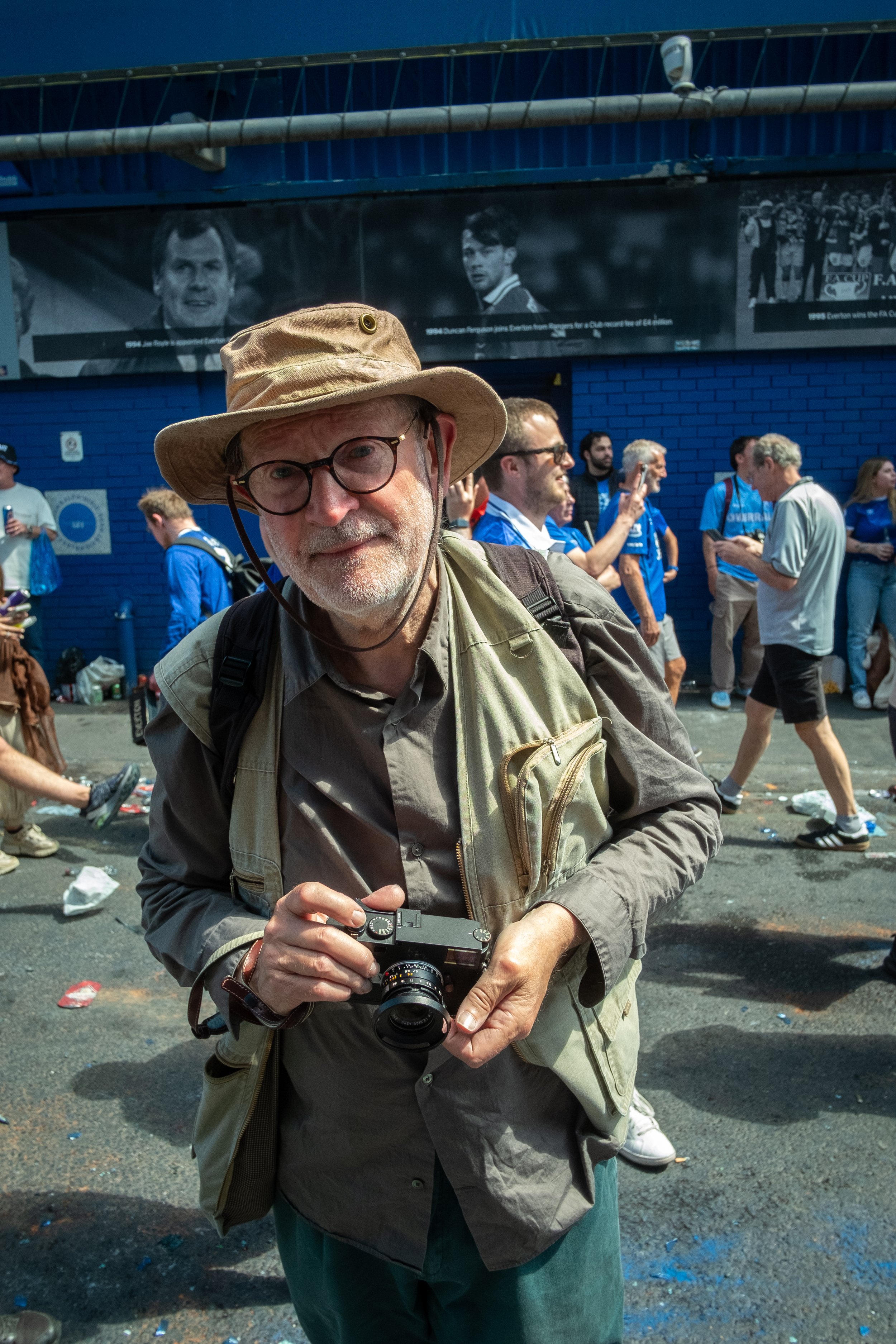 An older man with glasses, a beige hat, a beige vest, and a gray shirt holding a camera, standing among a crowd of people on a street with a blue wall in the background and a large black and white photos or advertisements above.