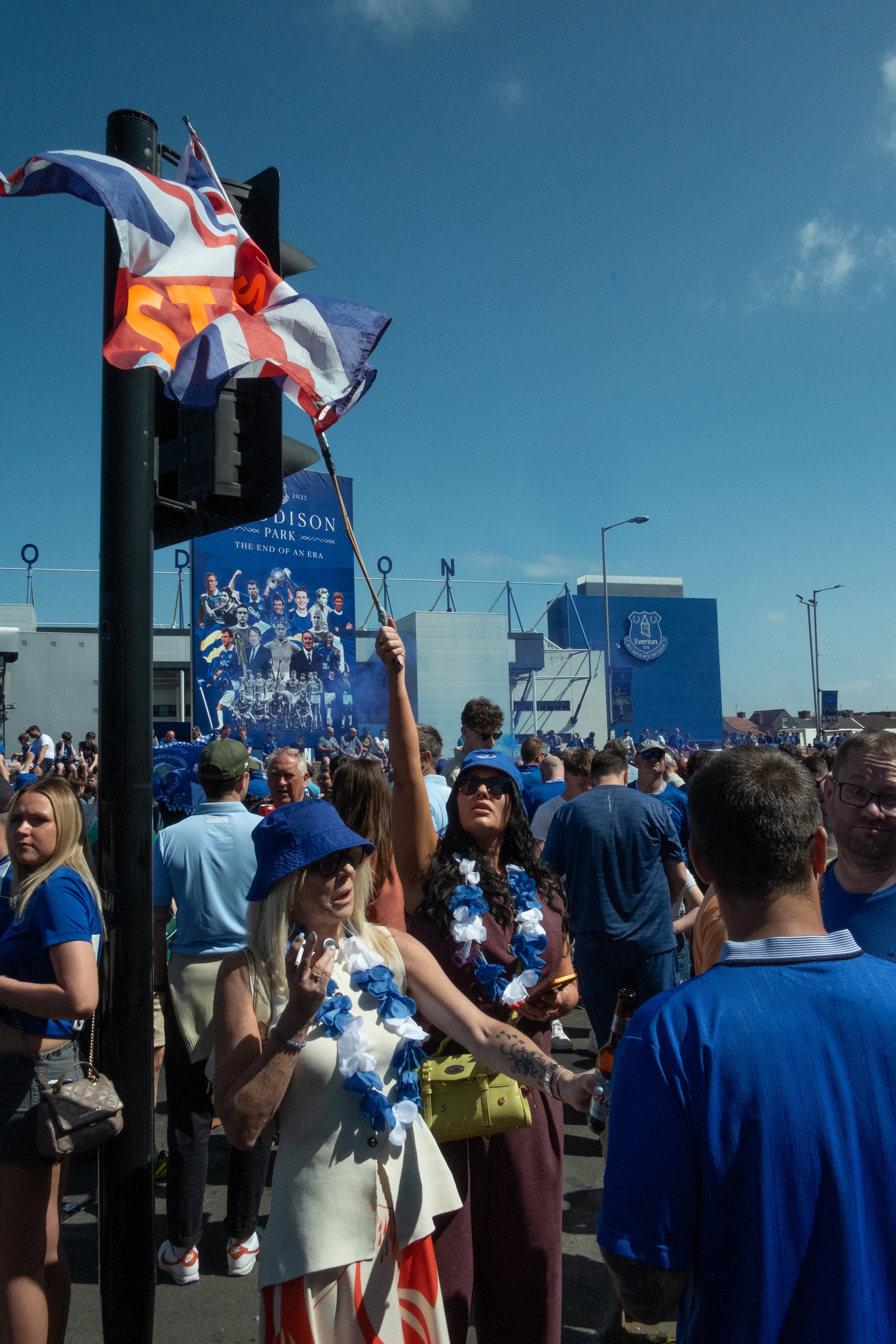 Crowd of people outside Everton football stadium with banners, flags, and fans in blue attire on a sunny day.
