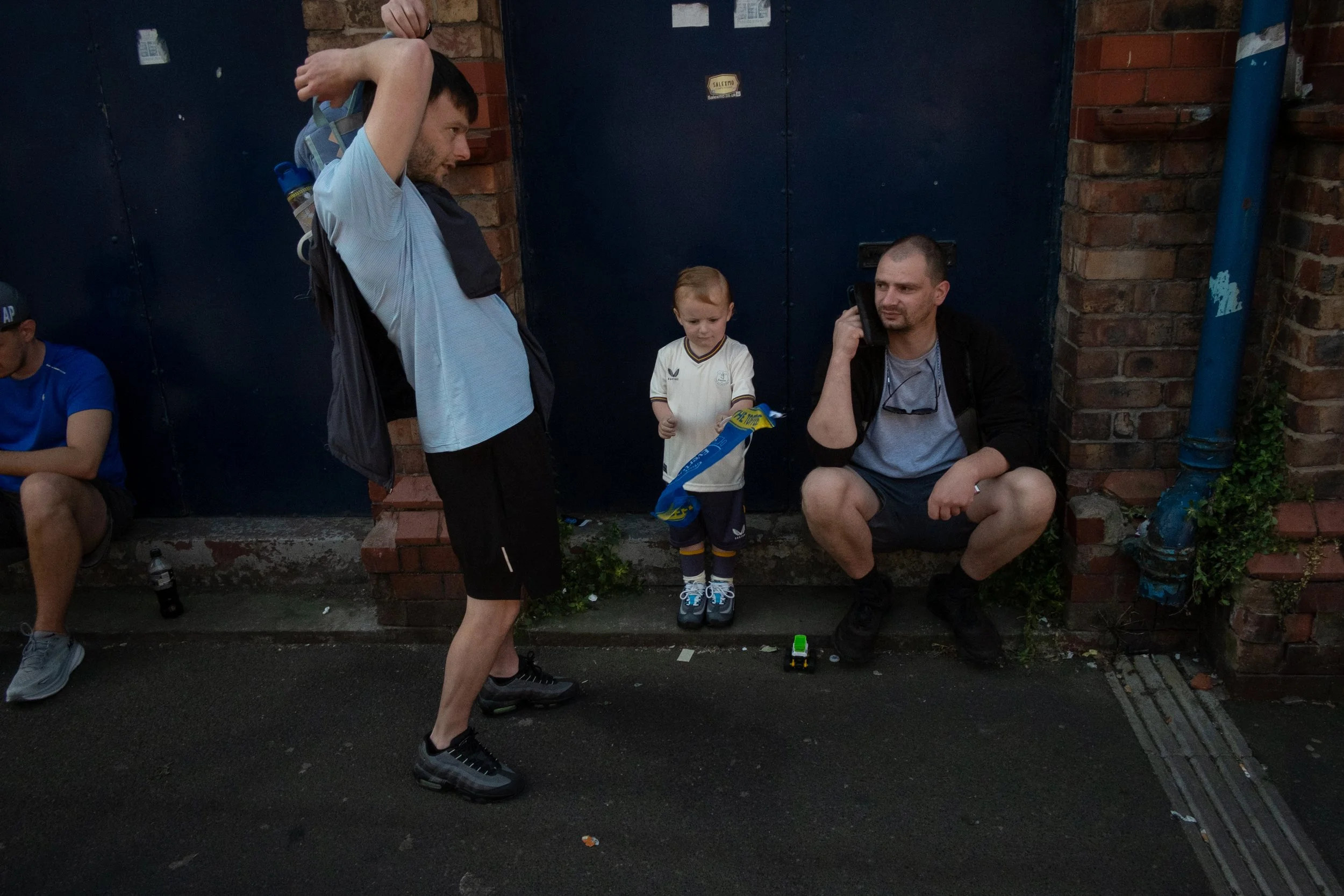A group of men and a young boy sitting and standing outside near a brick wall, some on the sidewalk and others on a ledge, with one man talking on a phone and the boy holding a blue and yellow item.