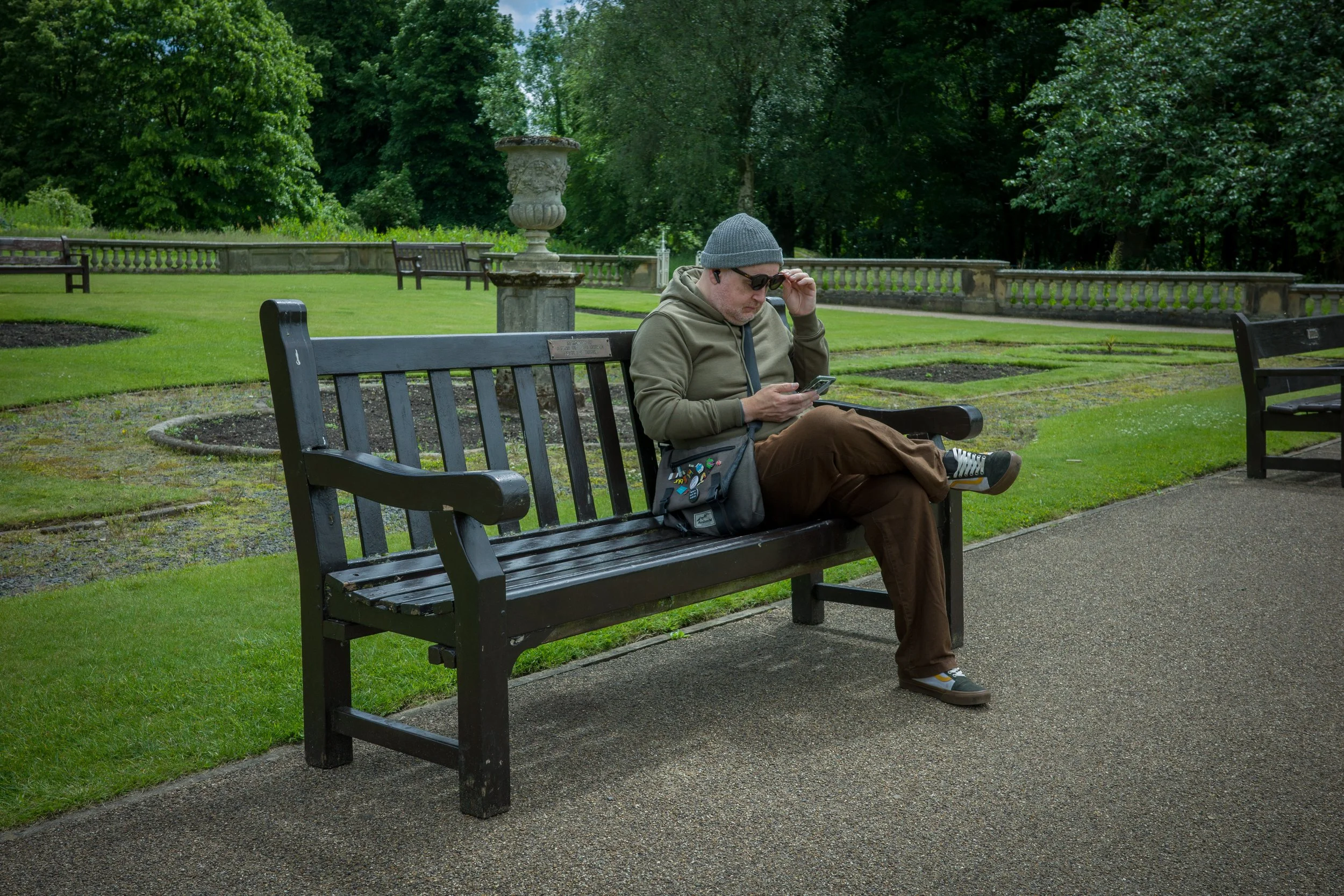 A man sitting on a park bench, wearing sunglasses, a gray beanie, a hoodie, and brown pants, looking at his phone. The park has green grass, trees, and decorative stone planters.