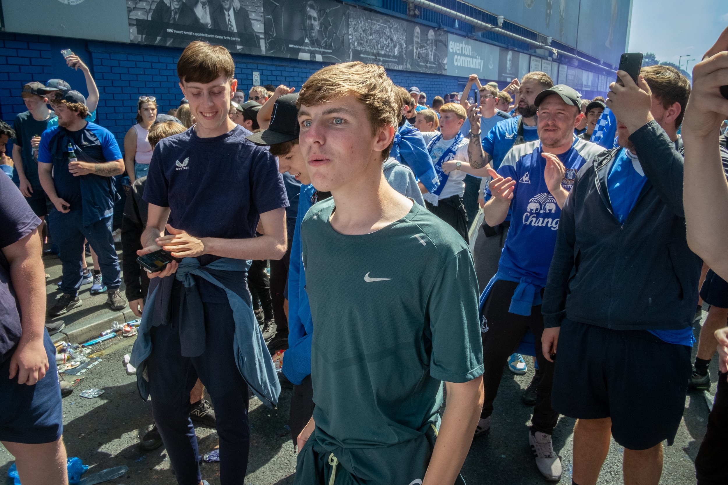 Young man in a green Nike shirt standing among a crowd of people dressed in blue, some clapping, others taking photos, outside on a sunny day near a blue wall with a large banner.