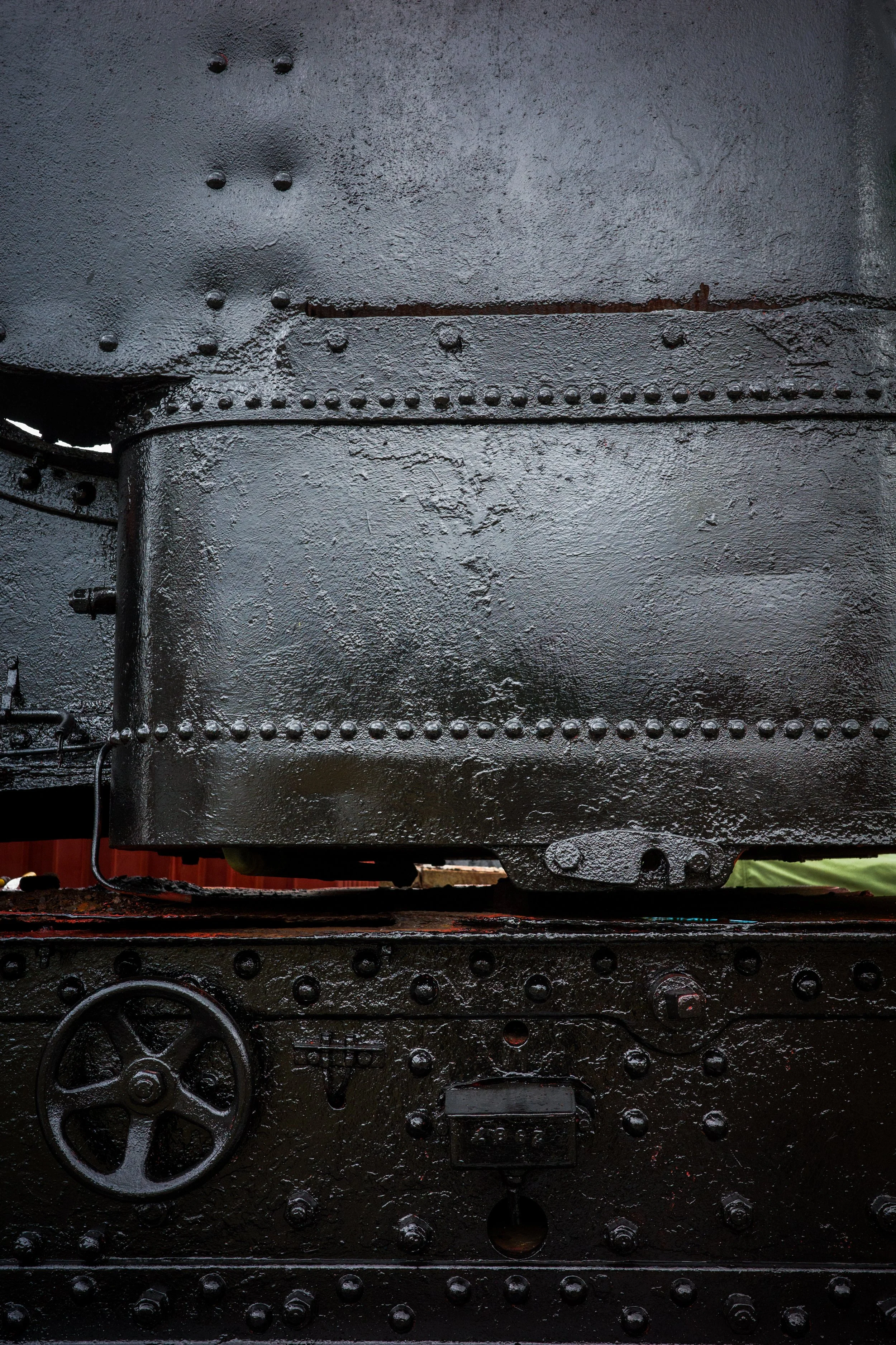 Close-up of a large, black iron machine with rivets, gears, and a wheel, likely part of an industrial or locomotive mechanism.