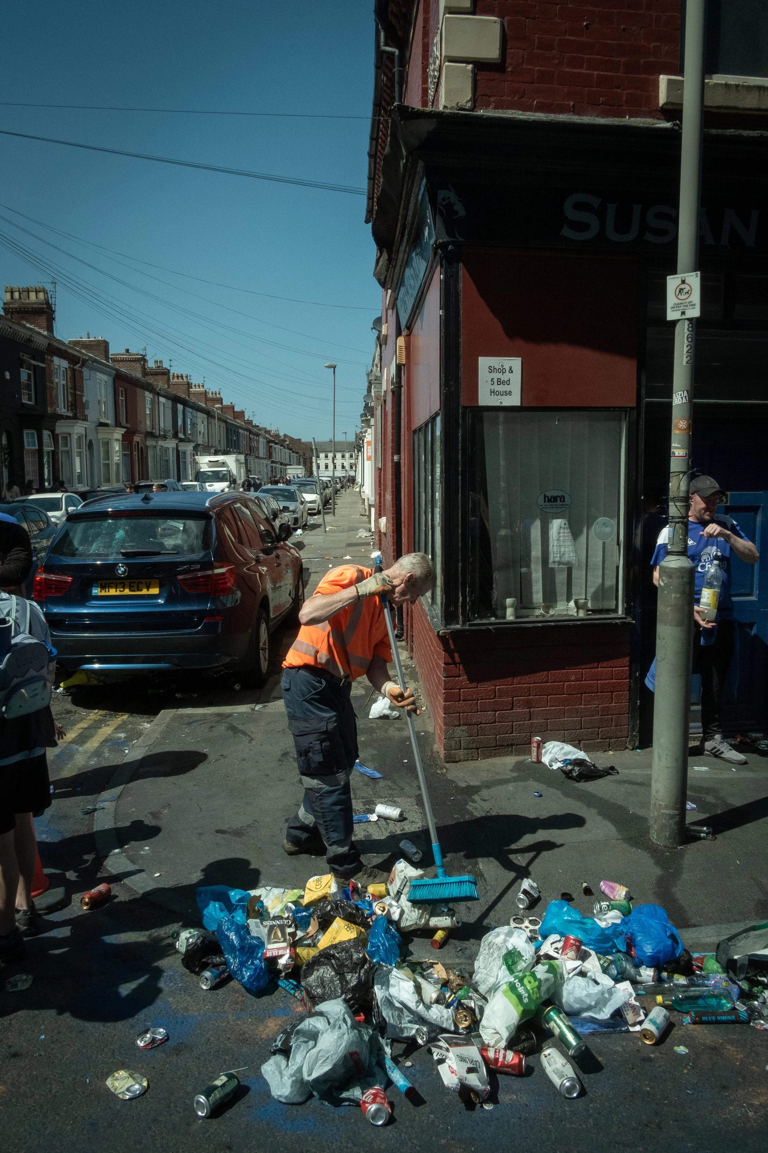 A man wearing an orange safety vest and blue pants sweeping trash, including cans and bags of garbage, on a city street with parked cars and row houses in the background.