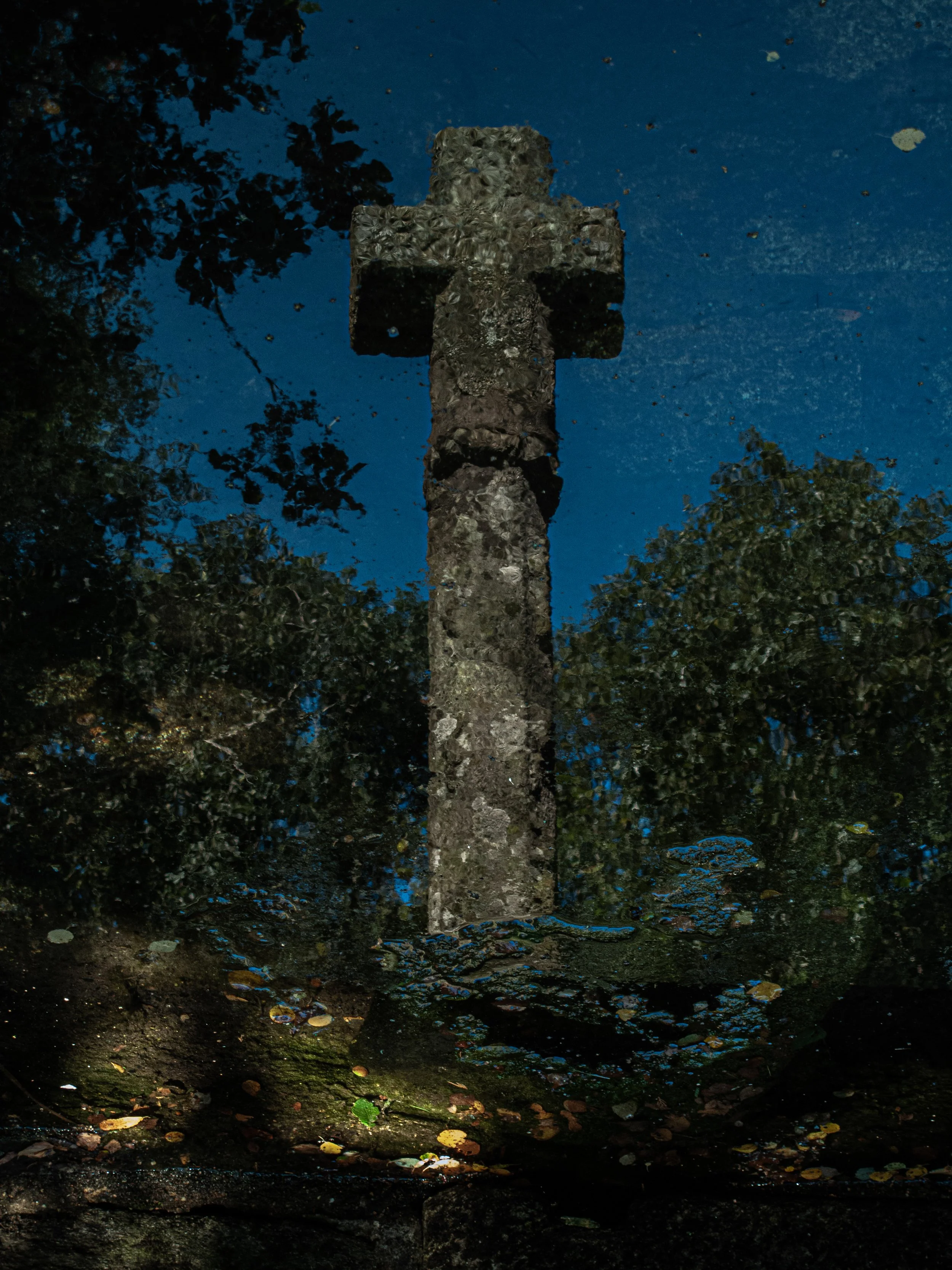 Reflection of a stone cross monument in a pond with trees and blue sky