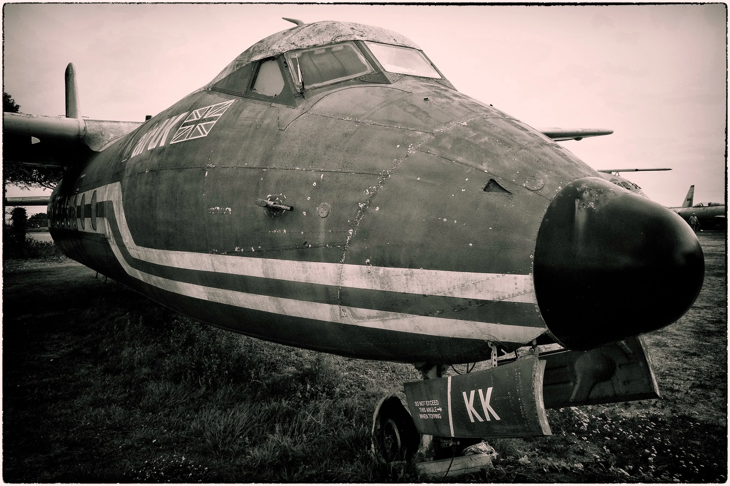 A large military aircraft on display outdoors, showing the front and cockpit section, with a Union Jack flag painted on the side, and weathered, damaged surface. There is a sign in front of it with regulatory information.