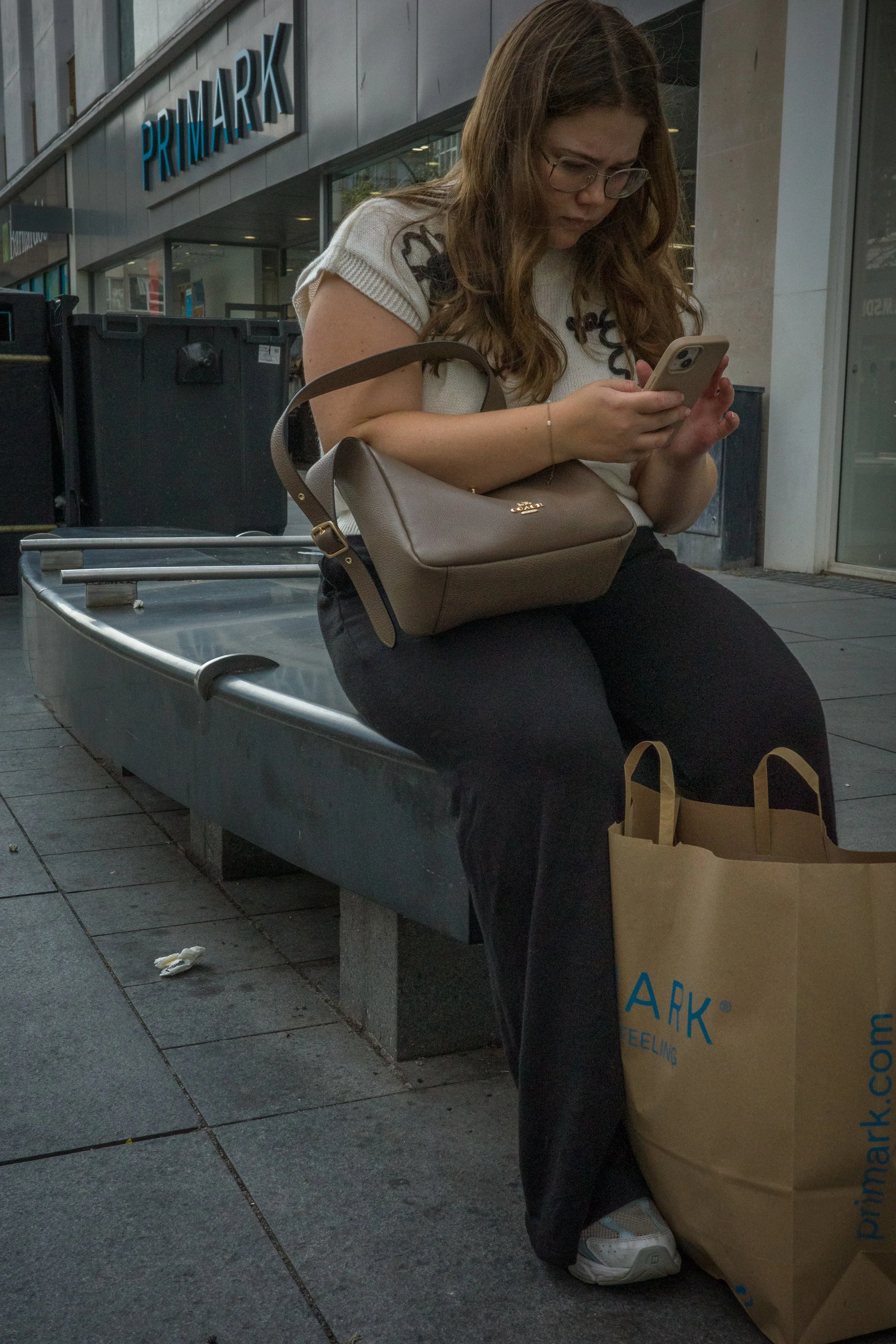 A woman wearing glasses and a white shirt sitting on a bank outside a Primark store, looking at her phone. She has a tan purse on her lap and a shopping bag beside her. There is a trashcan behind her and a trash piece on the ground.
