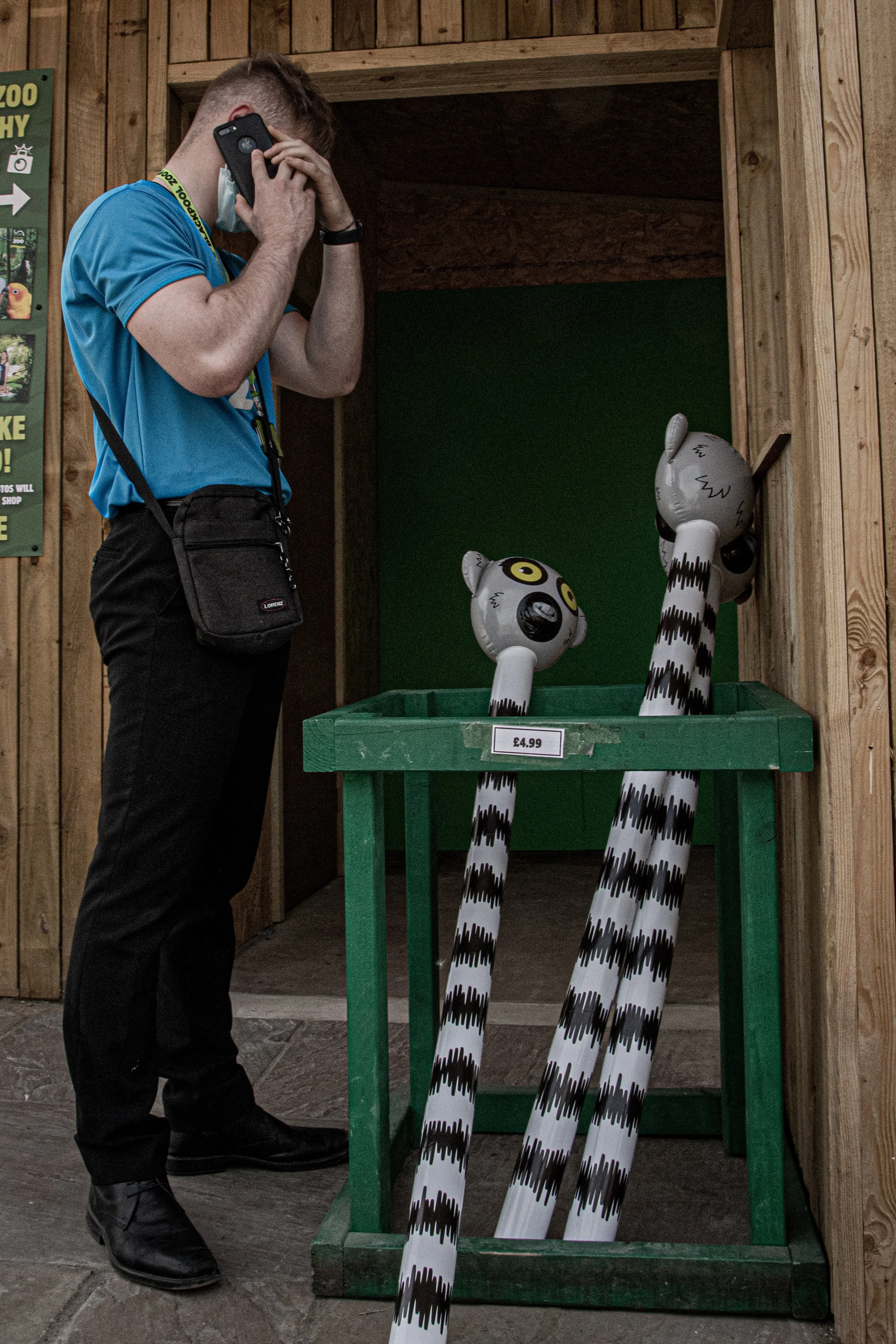 A man in a blue shirt and black pants standing inside a wooden enclosure, looking at and talking on a black cellphone. There are three tiger-striped inflatable objects with cartoonish tiger faces on a green table in front of him. The table has a pric