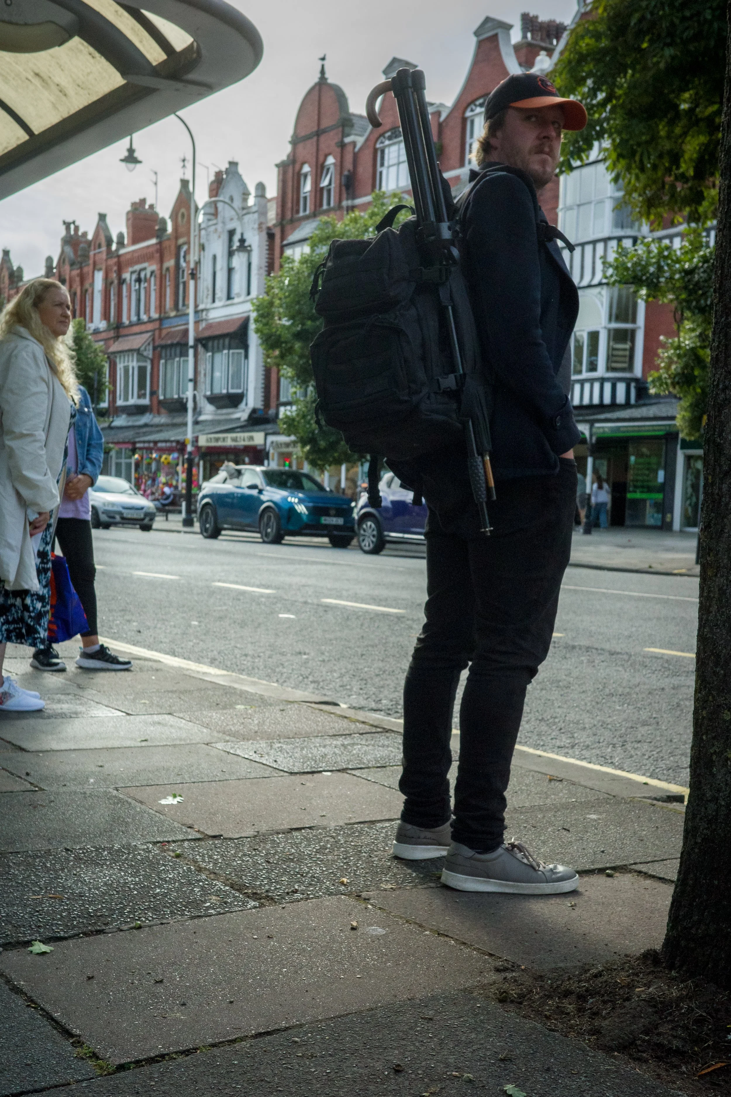 A young man standing on a city sidewalk, facing sideways with a backpack and a tripod strap over his shoulder. He wears a black jacket, black pants, gray sneakers, and a cap, with a row of colorful Victorian-style buildings across the street.