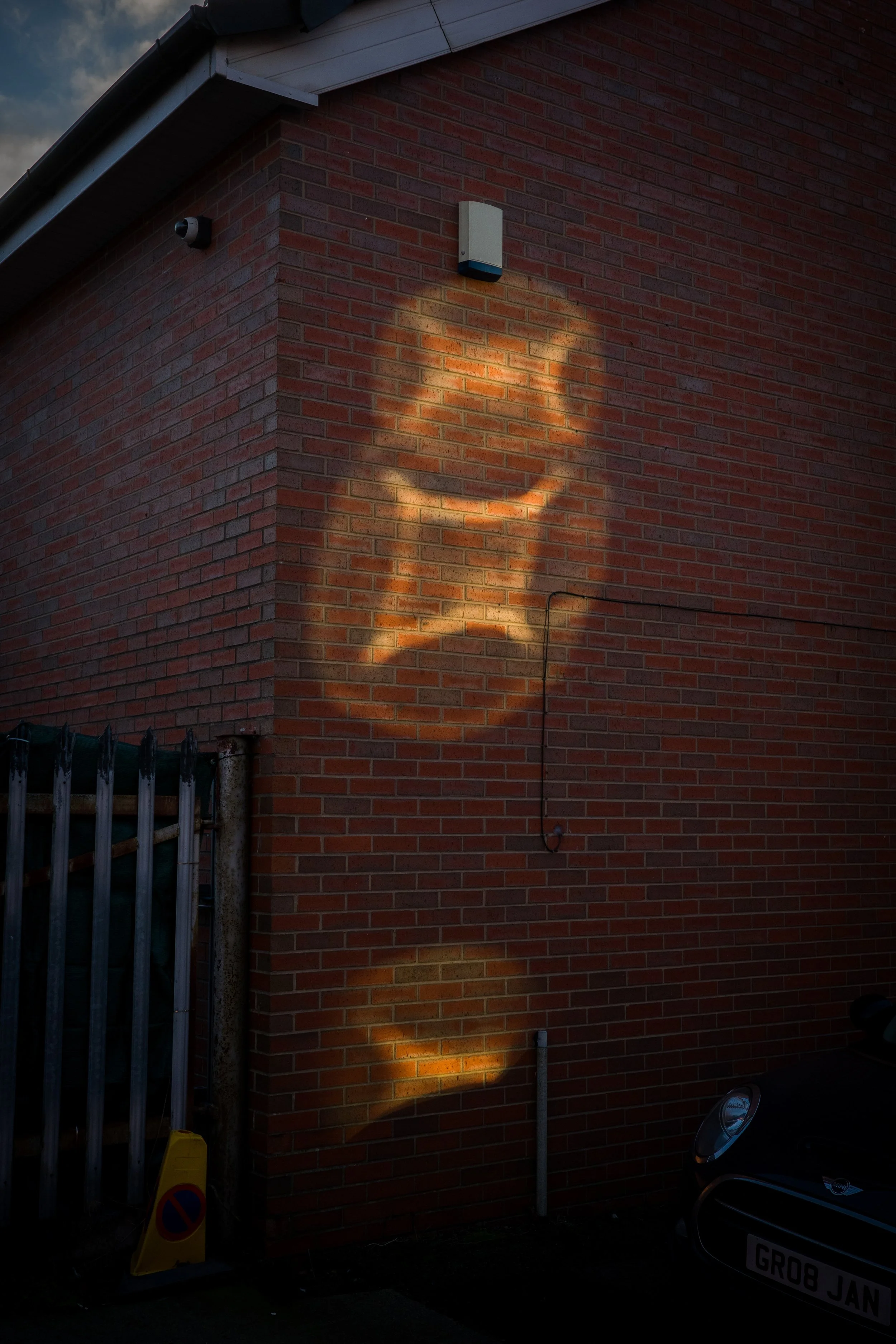 Brick wall of a building with a security camera, a motion sensor, a cable, a yellow 'no parking' sign, and part of a car with a visible license plate.