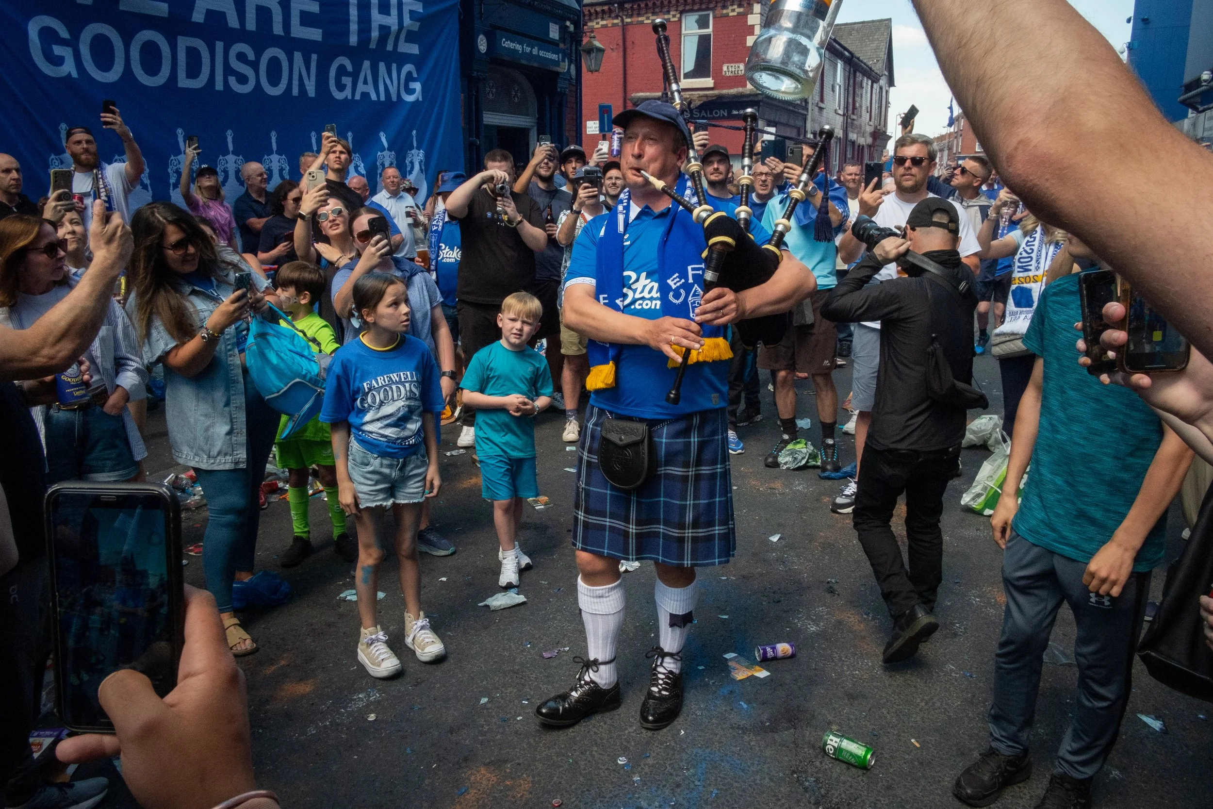 A man dressed in a traditional Scottish kilt playing bagpipes in the middle of a crowd during a street event. People around him are taking photos and videos, and the background shows banners and buildings.