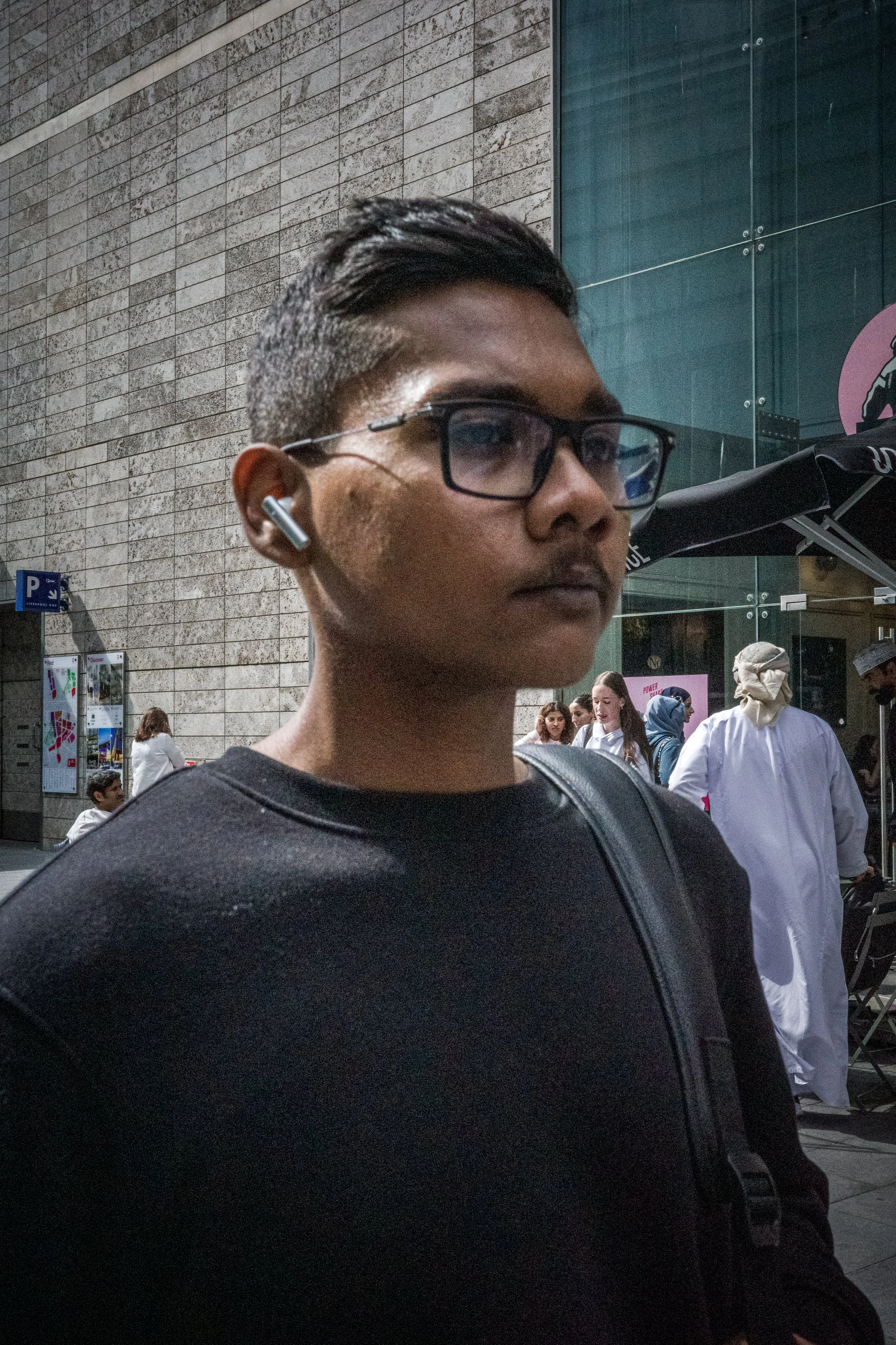 A young man with glasses and earbuds stands outdoors near a modern building with a stone wall and glass windows. Several people are visible in the background, including a person in a white medical coat.