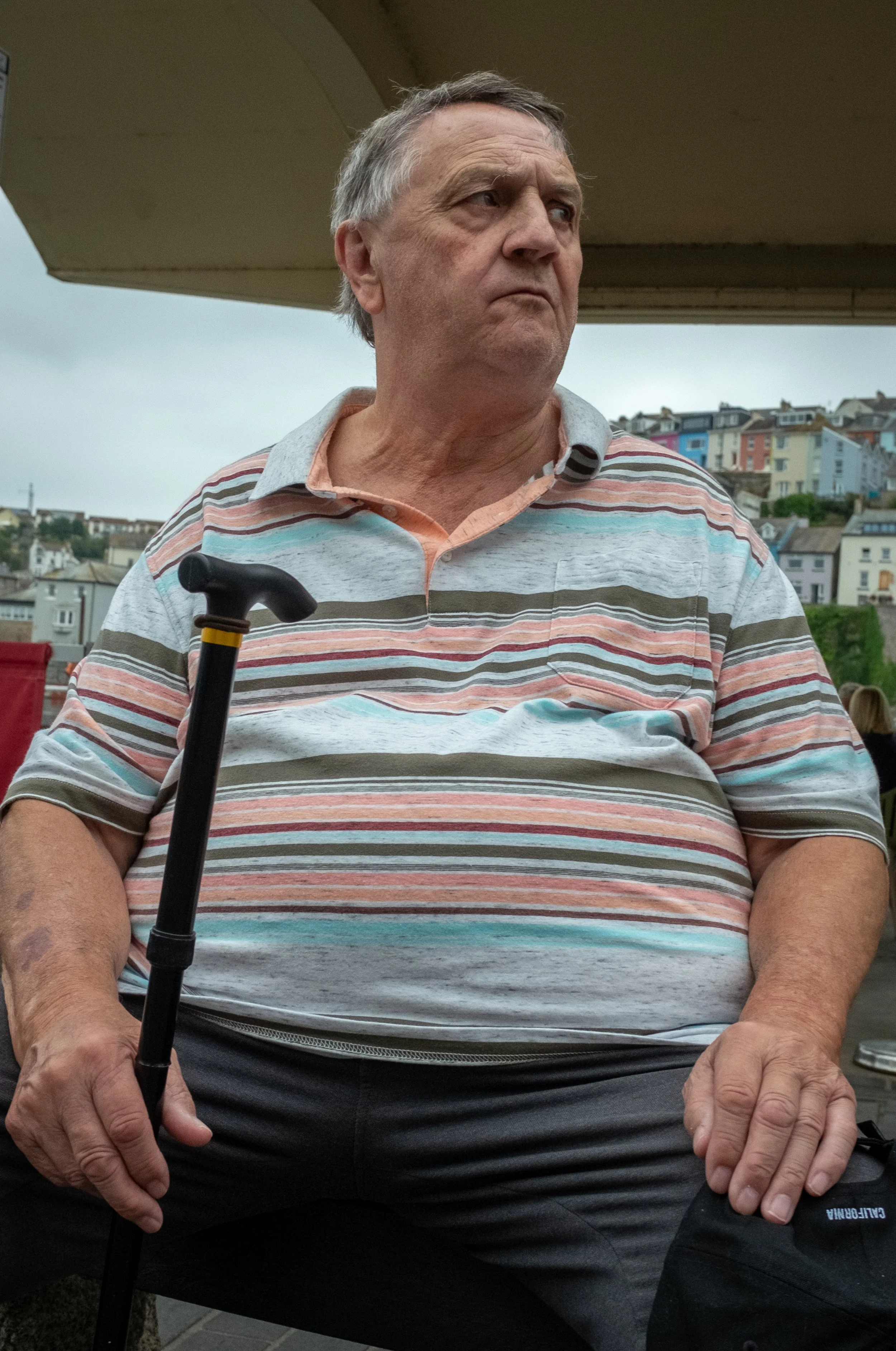 An older man sitting outdoors under a structure, holding a cane, wearing a striped polo shirt, with a hilly neighborhood and overcast sky in the background.
