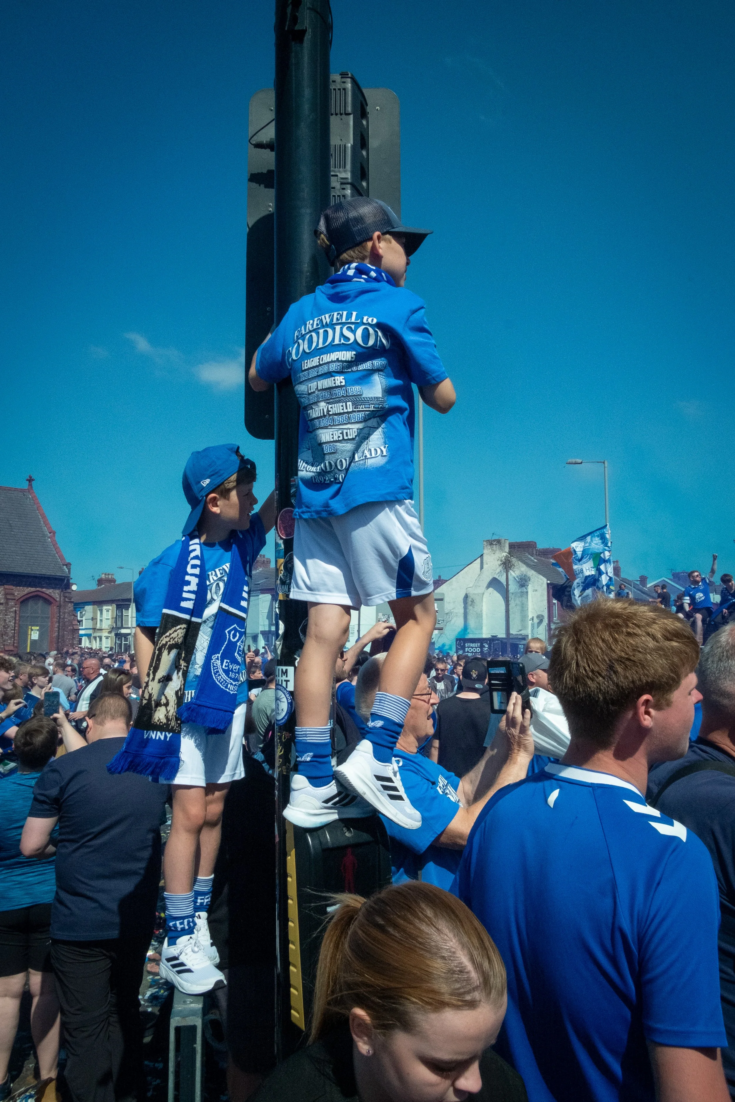Two young boys dressed in blue football jerseys and scarves standing on a crowd at a sports rally. One boy is standing on a platform, and the other is standing on someone's shoulders, surrounded by many people in blue jerseys, with flags and banners 
