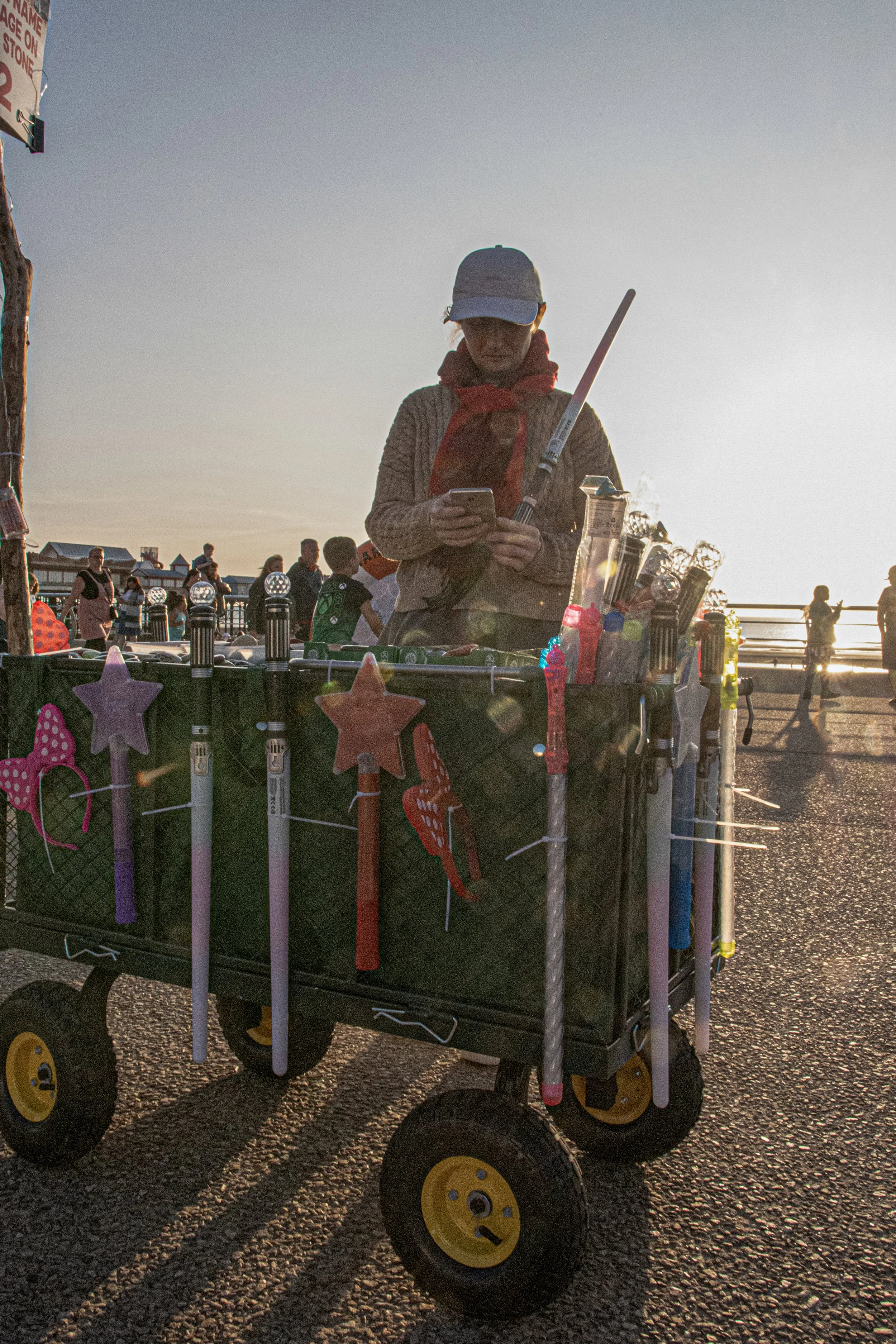 A person wearing a gray hat, red bandana, and beige sweater is looking at a smartphone while standing behind a carnival game cart with toy water guns, stars, and other prizes, at sunset outdoor event with people in the background.
