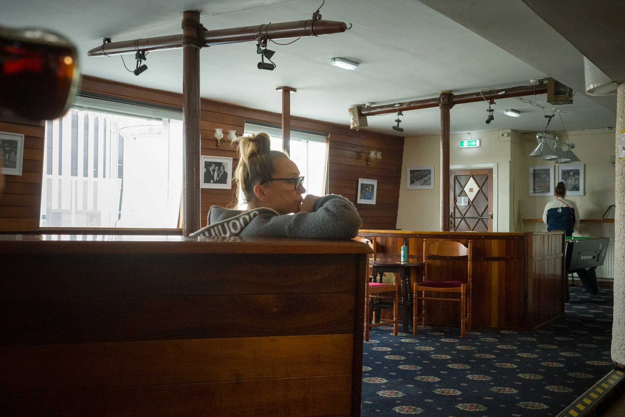 A woman with glasses and blonde hair tied in a bun, sitting alone at a booth in a cozy pub or cafe, looking to the right. The interior features wooden paneling, framed photographs on the walls, and a pool table in the background. Sunlight streams thr