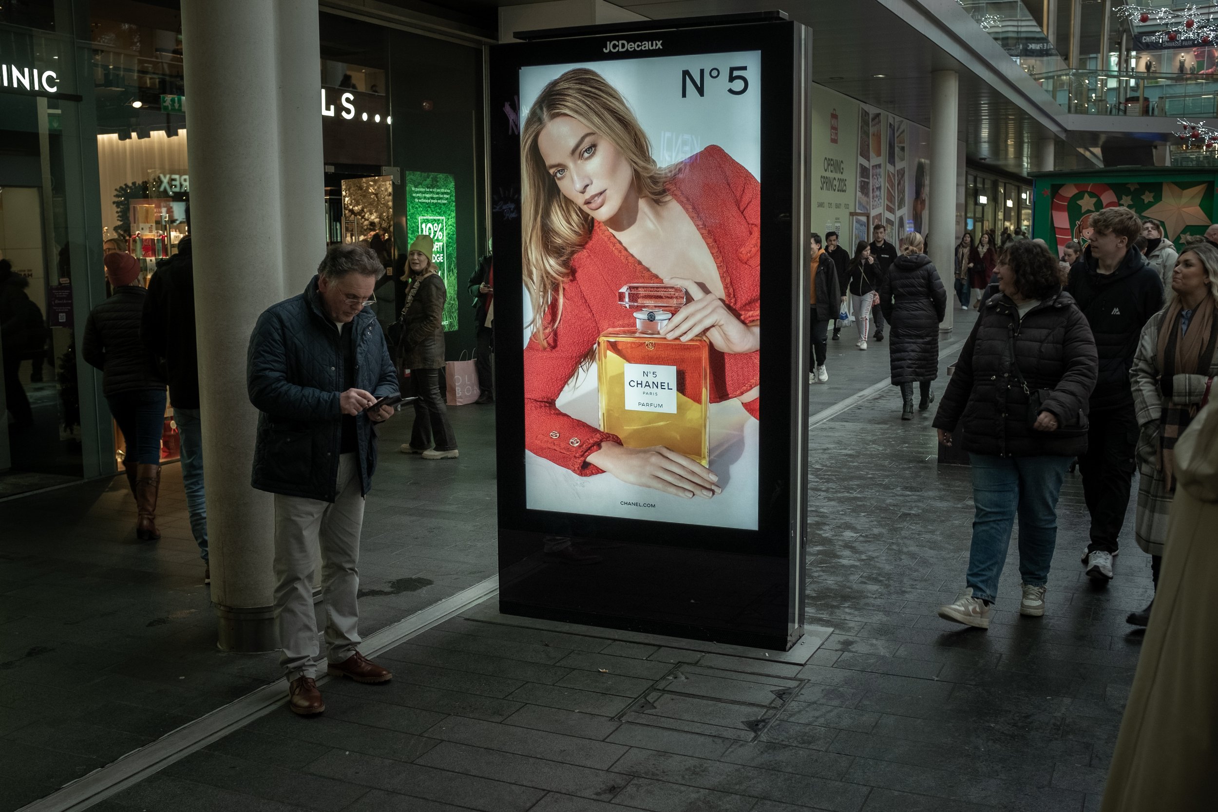 A busy shopping mall with a large advertisement for Chanel No. 5 perfume featuring a woman in a red sweater holding a perfume bottle. People are walking around, some looking at their phones, near stores and signs, including a Christmas-themed display