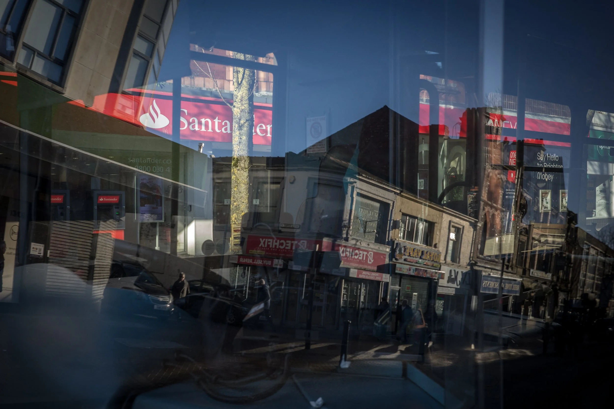 A street scene with storefronts, cars, and pedestrians, reflected in a large glass window.