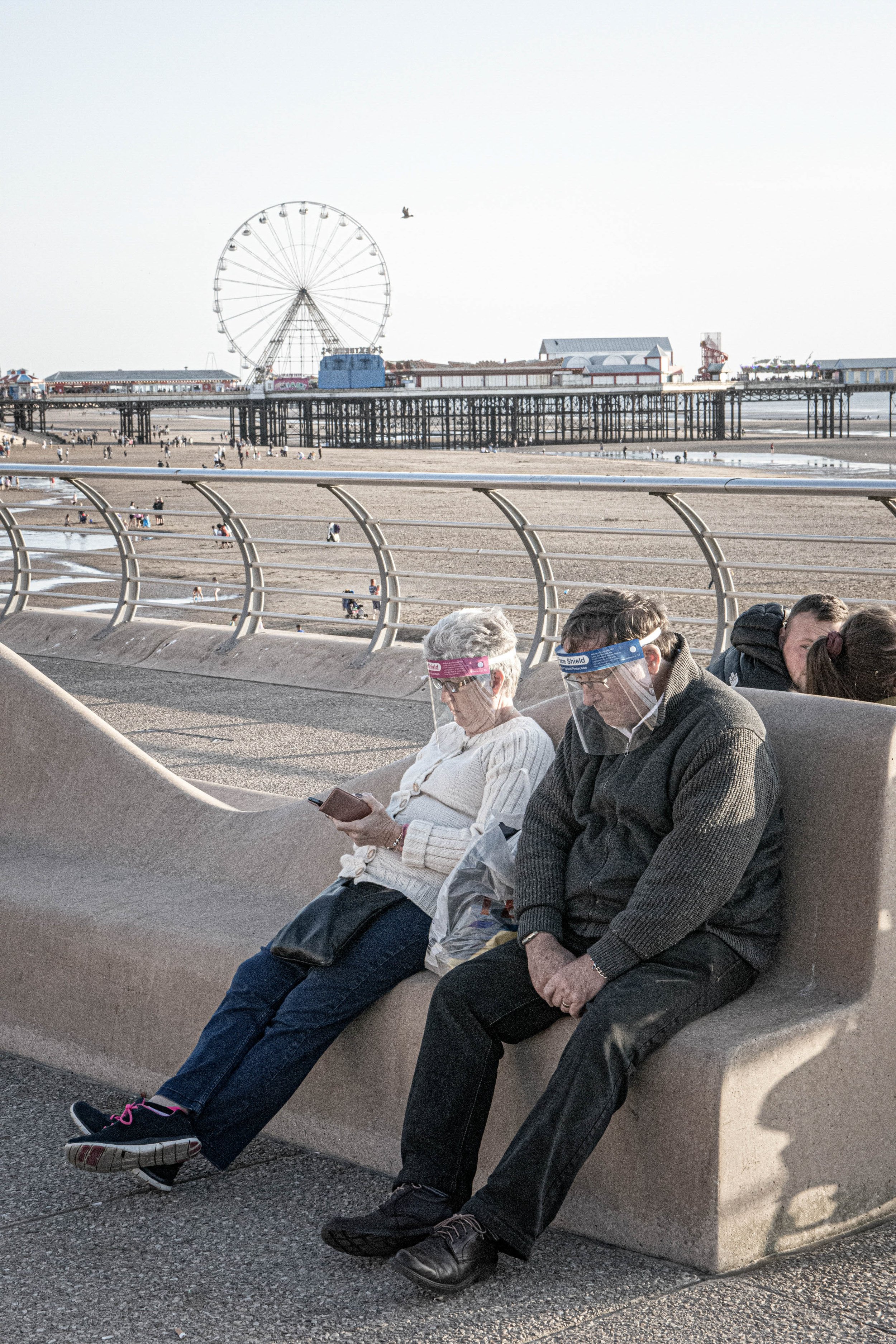 People sitting on a concrete bench near the beach, with an amusement park pier and Ferris wheel in the background.