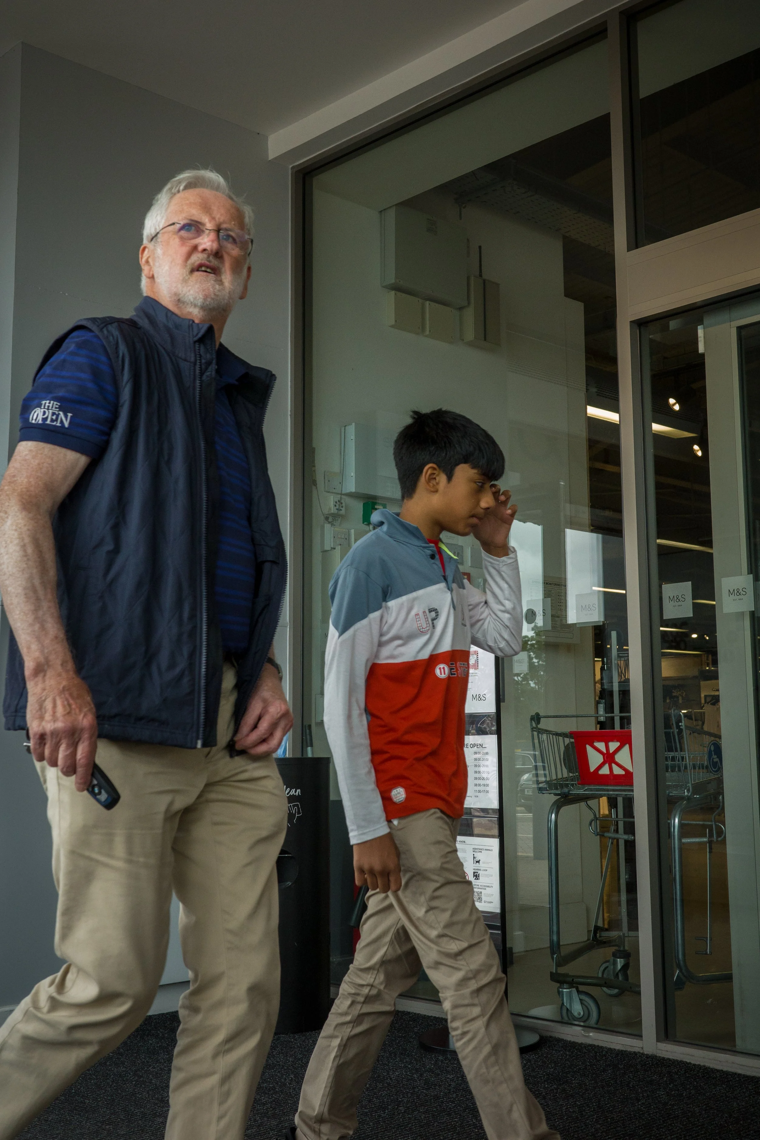 An older man with gray hair and a beard wearing glasses, a navy vest, and beige pants walks past a young boy with dark hair wearing a red, white, and gray jacket and tan pants in front of a glass door marked 'M&S' at a shopping center.